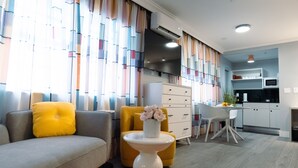 Ein modernes Wohnzimmer mit einer Couch, einem gelben Sessel und einem weißen Couchtisch mit Blumen. Eine Küchenzeile mit Mikrowelle und ein Esstisch mit Stühlen sind ebenfalls vorhanden.