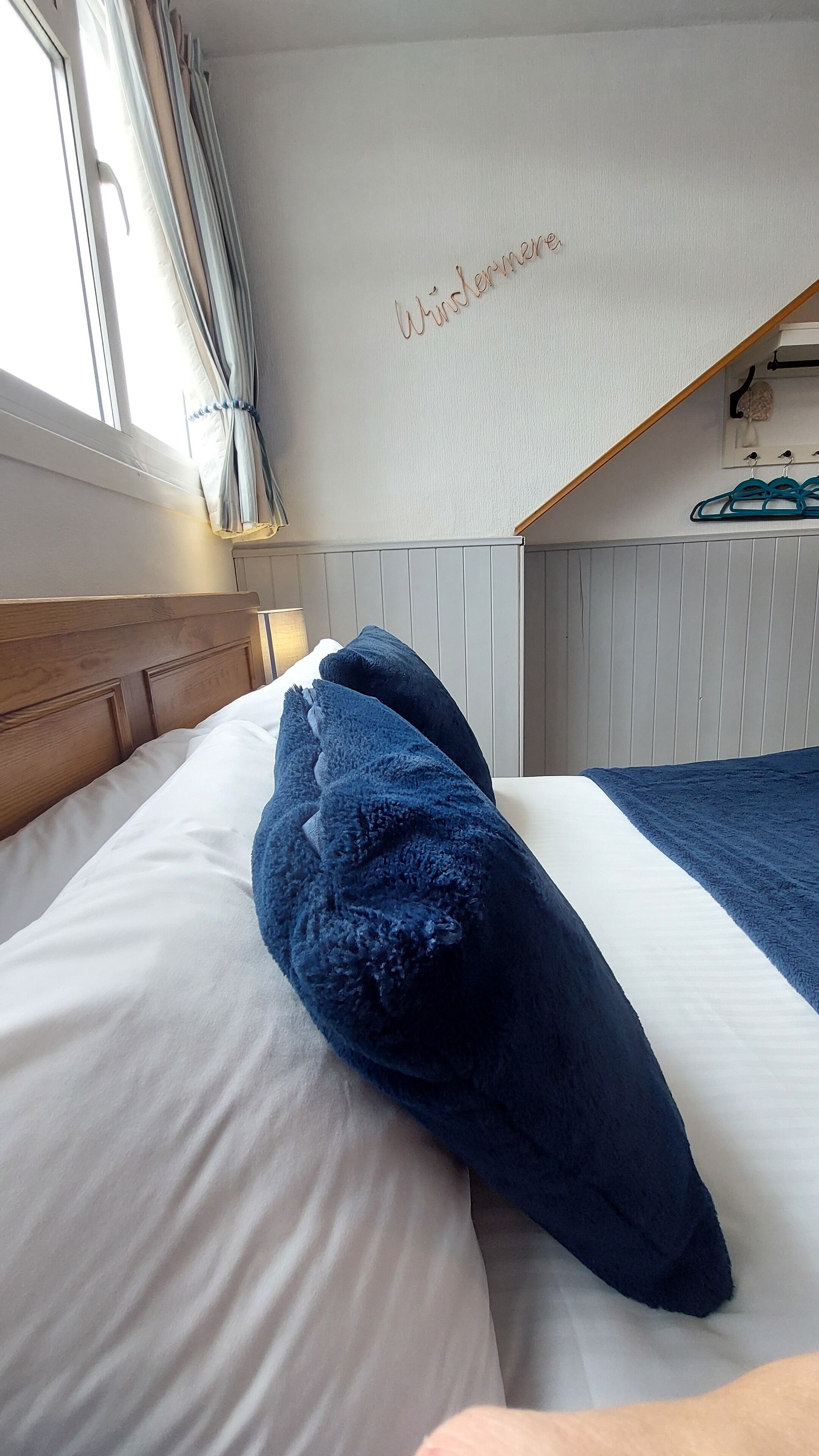 Una habitación de hotel con una cama, un ventanal con cortinas, una pared con un nombre y un perchero con ganchos.
