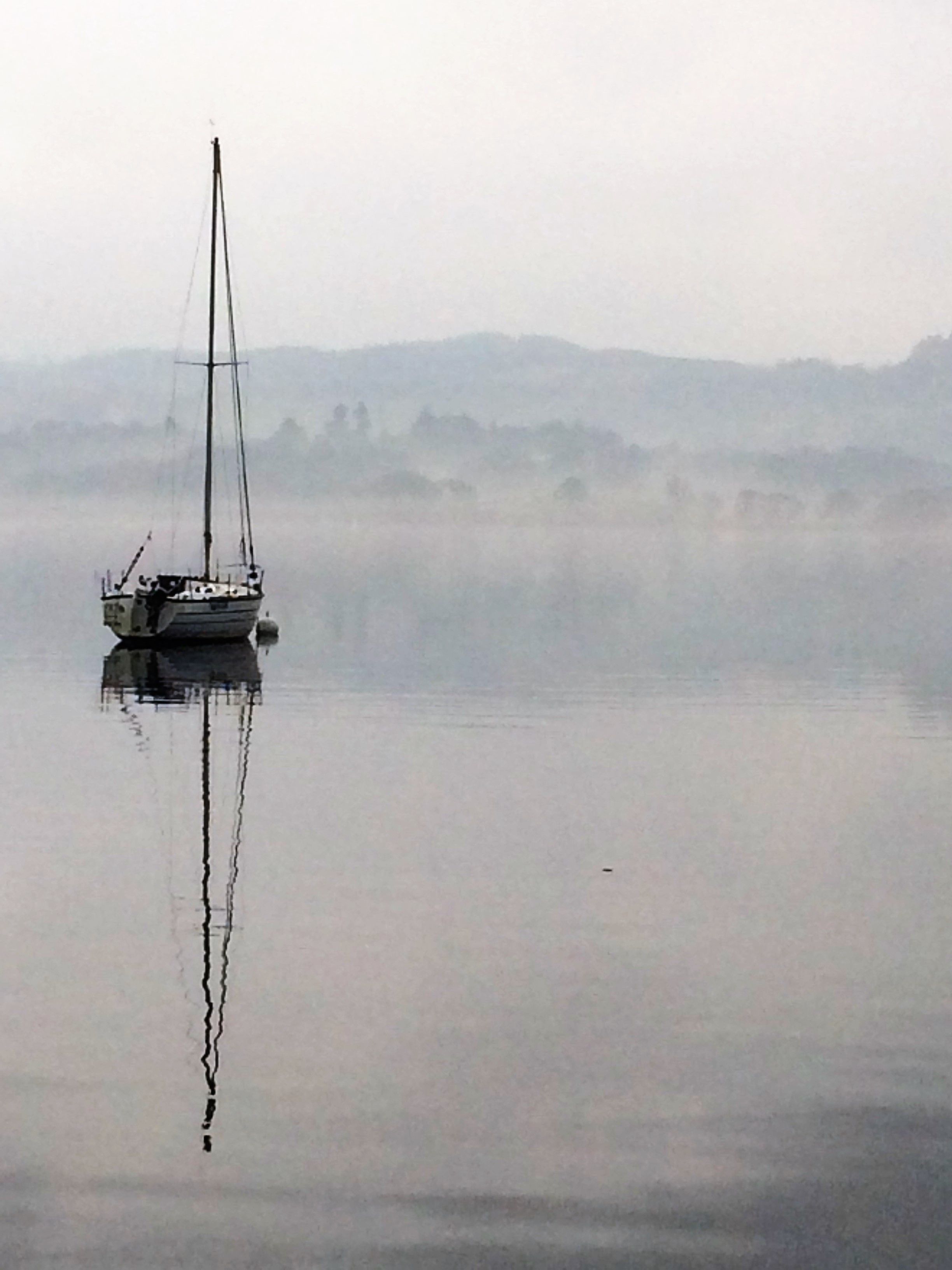 Un velero sobre aguas tranquilas, con un fondo brumoso y colinas lejanas.