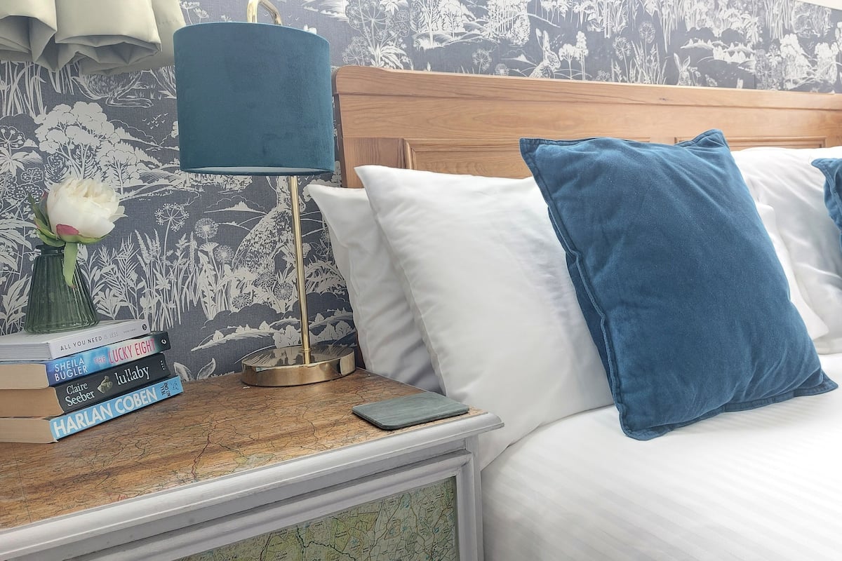 Ein ordentlich bezogenes Bett mit weißen und blauen Kissen, ein Nachttisch mit Lampe, ein Bücherstapel und eine Karte.
