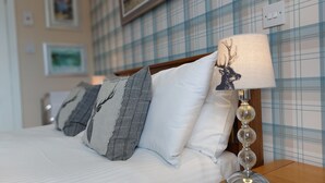 A neatly made bed with white bedding and decorative pillows, a bedside table with a lamp, and a plaid-patterned wall. - Invergarry Room Only Guest House (Windermere)