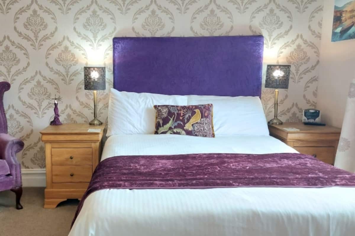 Ein Schlafzimmer mit einem großen Bett, einem violetten Sessel, zwei Nachttischen mit Lampen und einer Wand mit Muster.