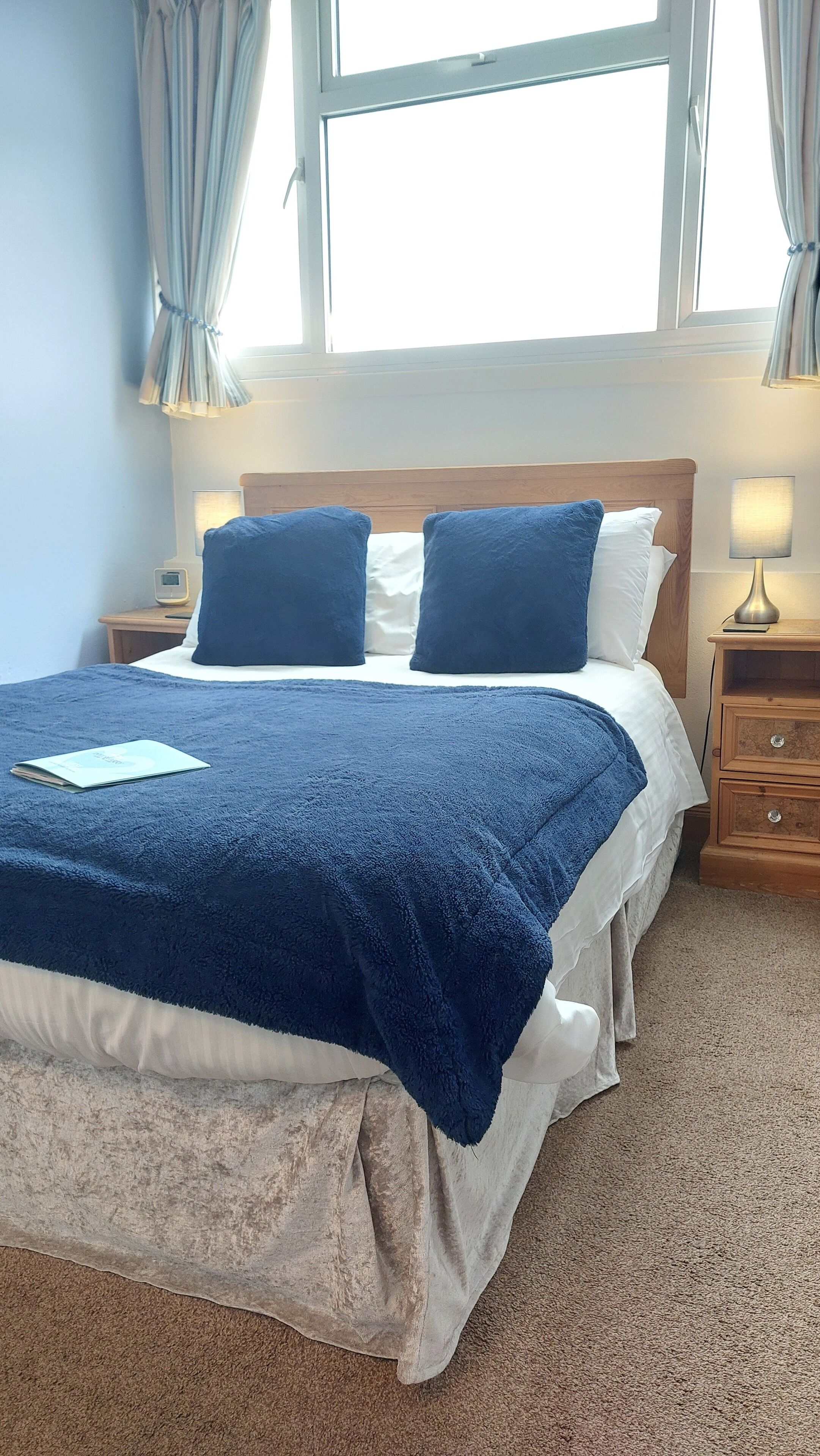 A neatly made bed with a blue blanket and pillows, a bedside lamp, and a nightstand with a book.