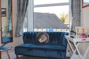 Un sofá azul con un cojín decorativo, una mesita con una cafetera y una ventana con cortinas.