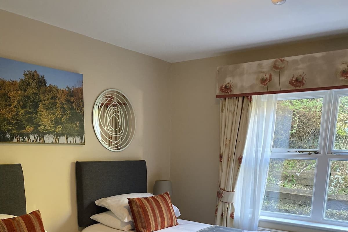 A hotel room with two beds, a large window with curtains, a painting on the wall, and a ceiling light.