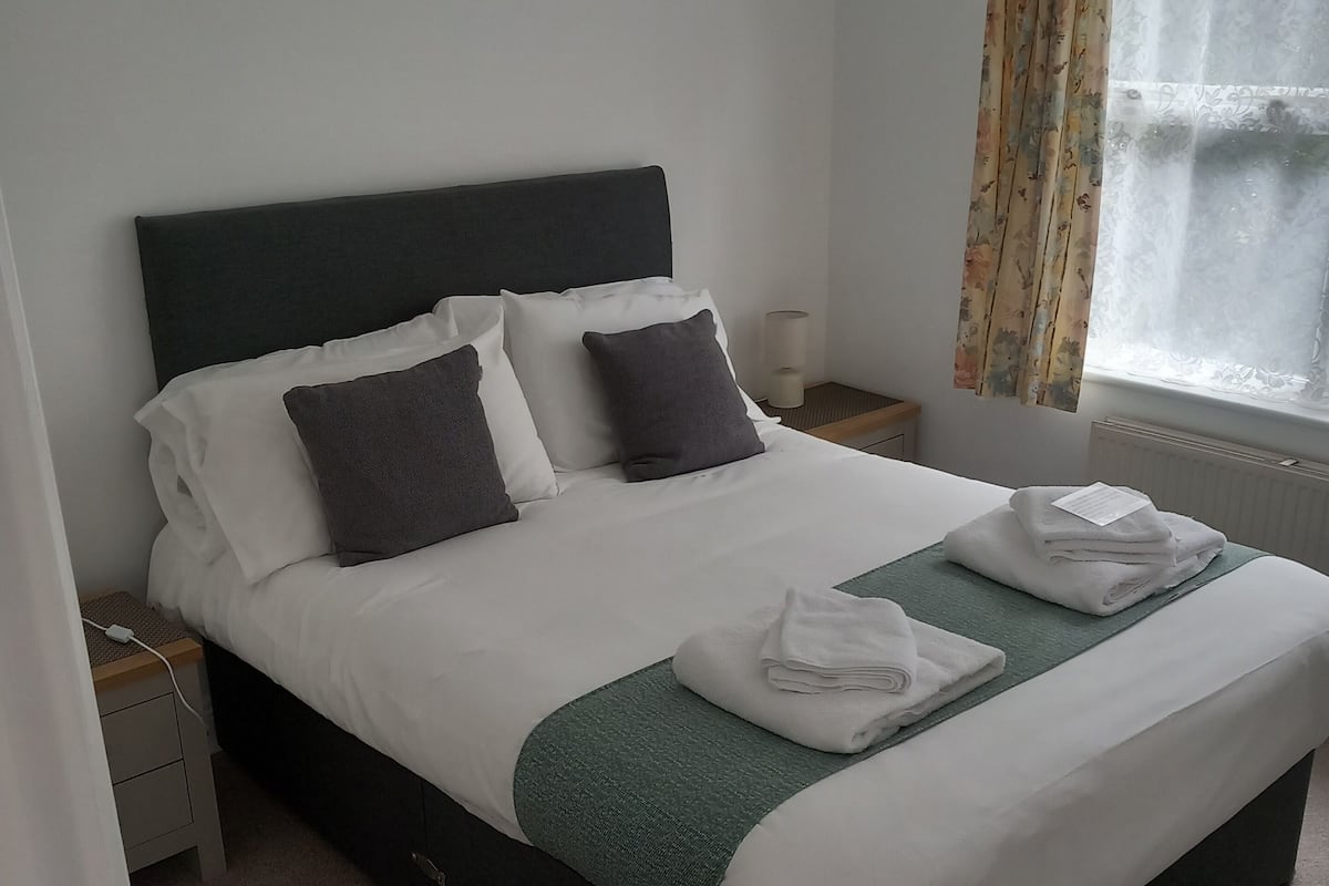 A neatly made bed with white and dark grey pillows, a green throw blanket, and folded towels on a wooden nightstand.
