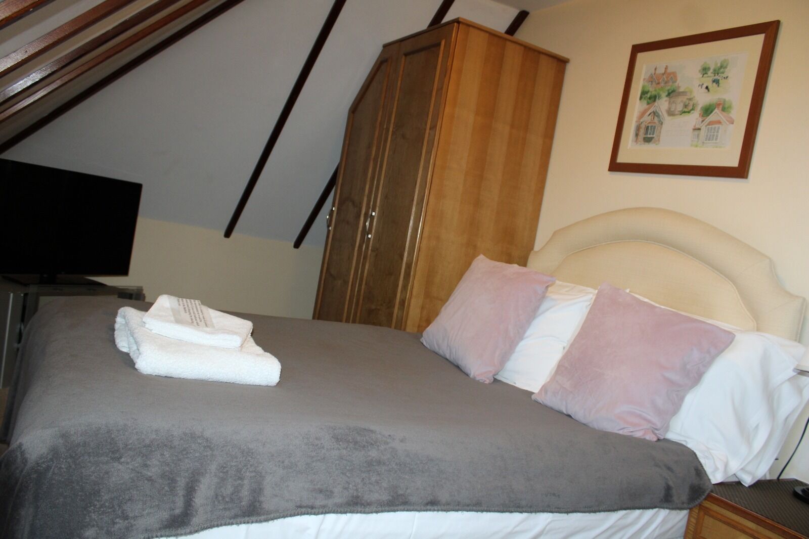 A bedroom with a bed, pillows, a blanket, a wooden wardrobe, and a framed picture on the wall.