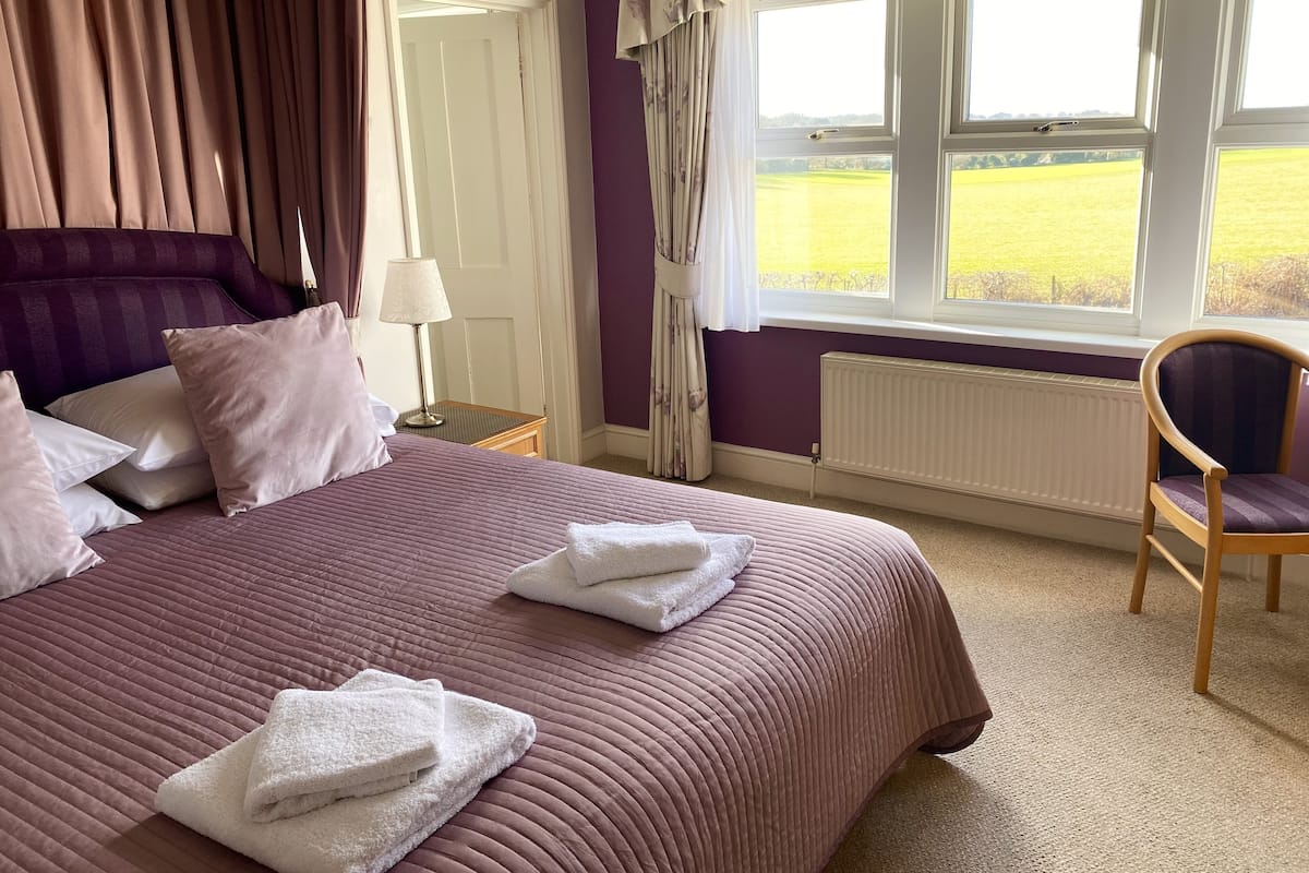 A neatly made bed with pillows and towels, a chair, a nightstand with a lamp, and a window with a view of a field.