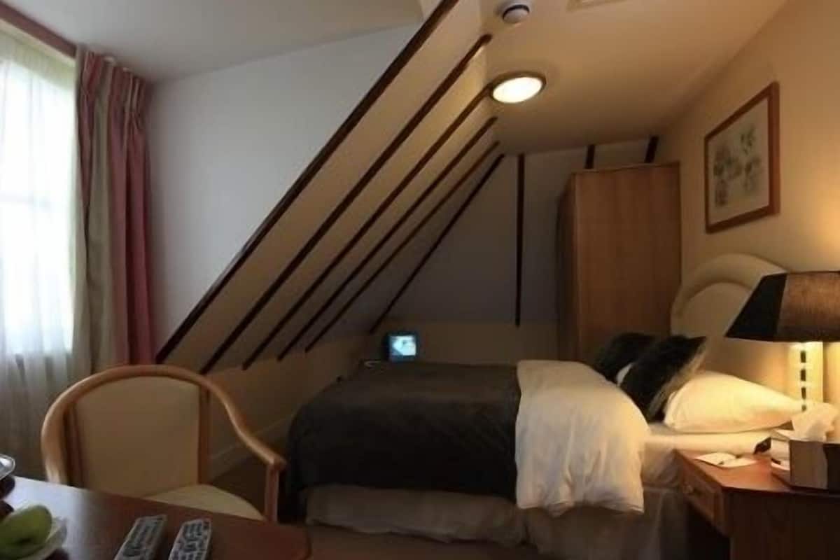 A compact hotel room with a bed, a desk, a chair, a lamp, and a TV.