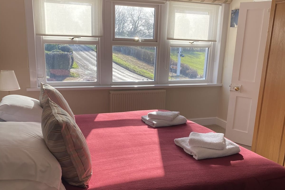 A bedroom with a bed, pillows, a window with a view of a road and greenery, and a radiator.