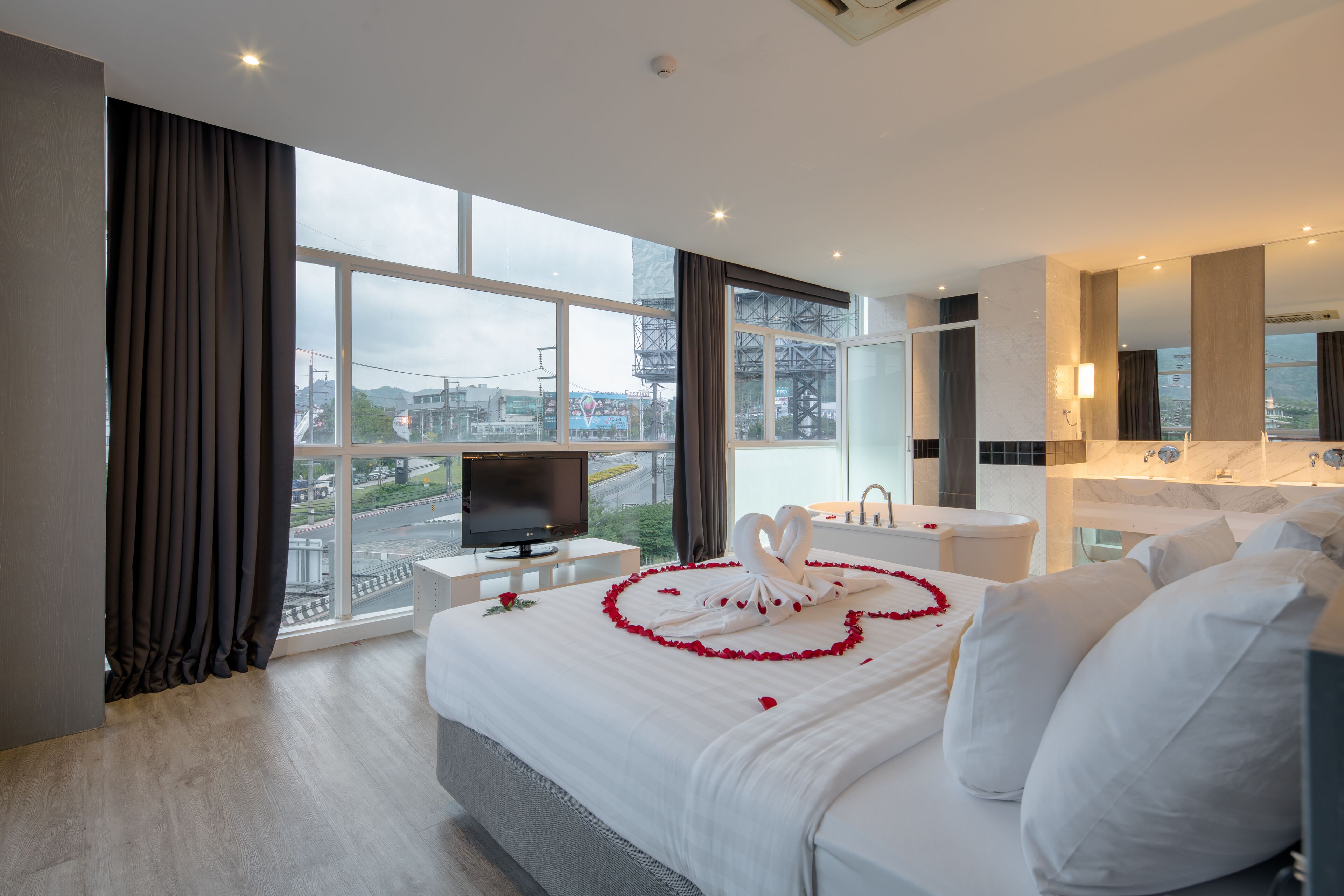 A modern hotel room with a large bed, a television, a bathroom with a bathtub, and a view of the cityscape.