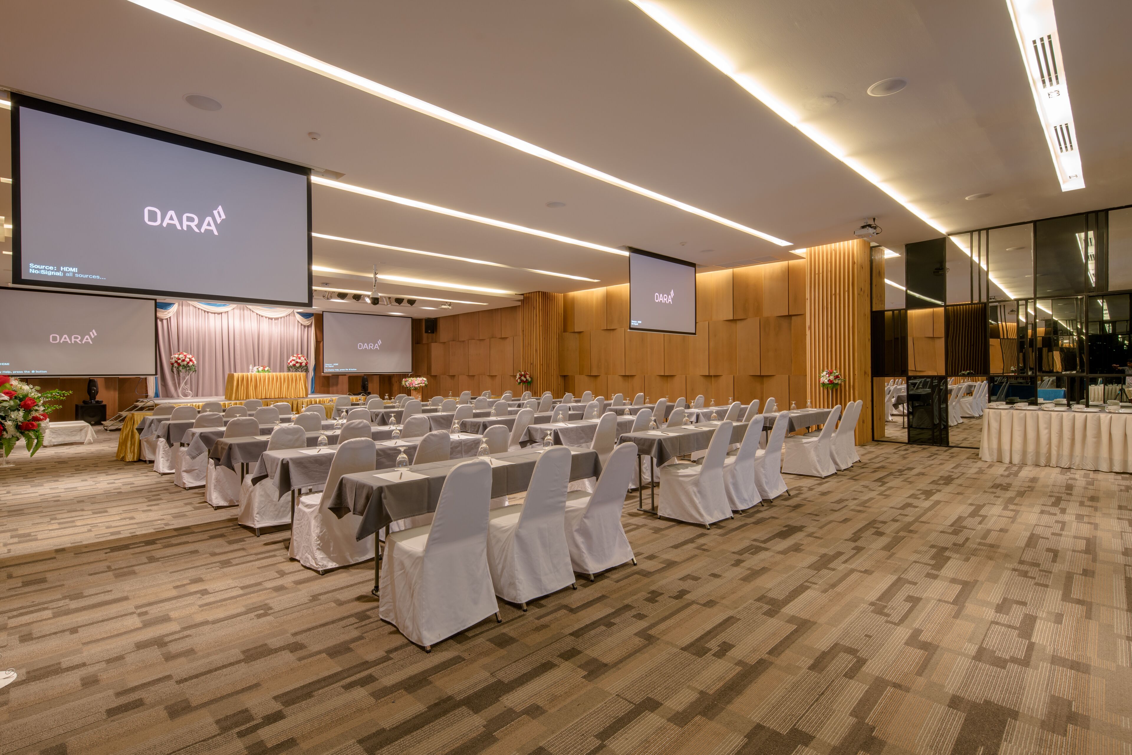 A conference room with rows of tables and chairs, large screens displaying 'DARA', and a carpeted floor.