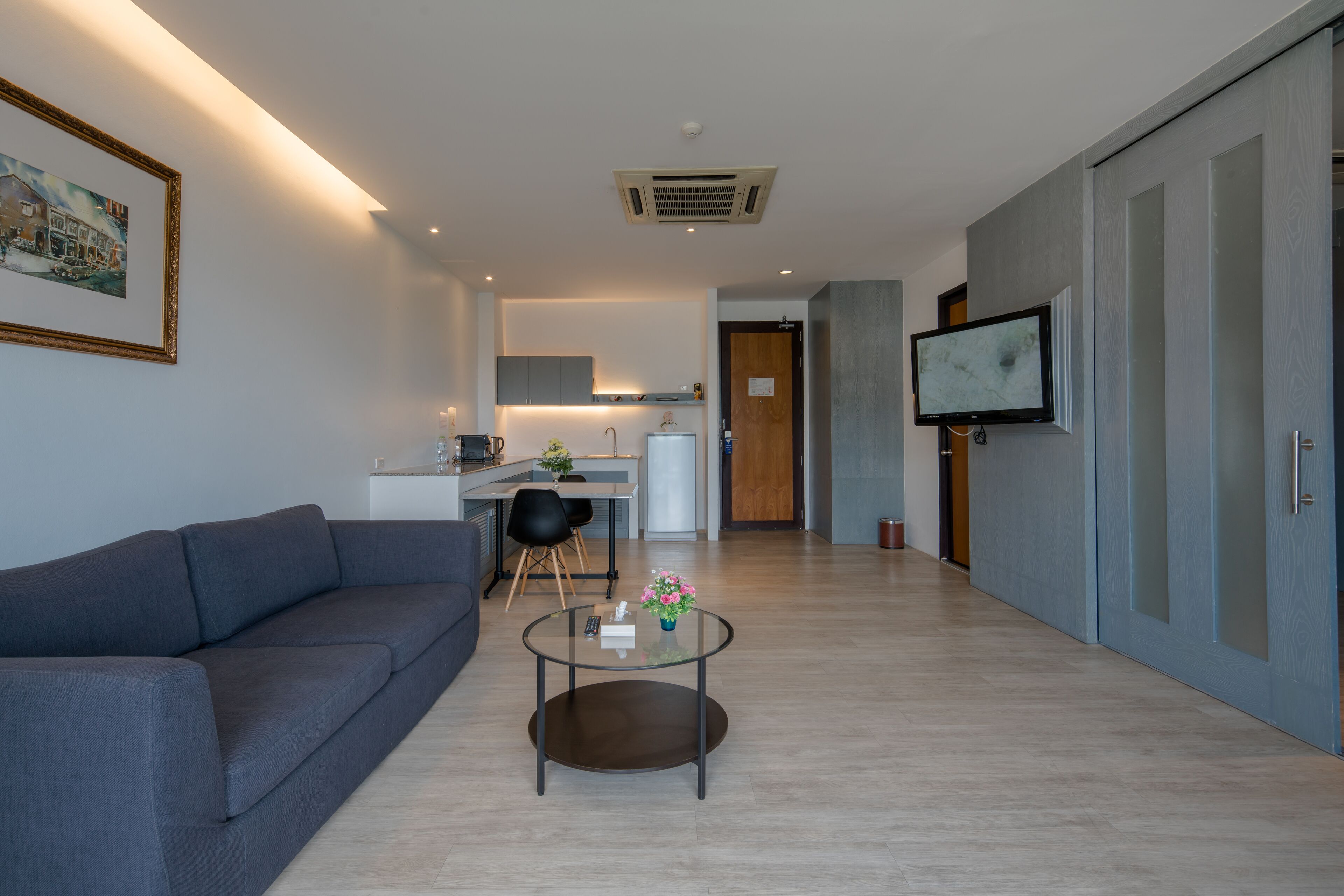 Ein modernes Wohnzimmer mit einem grauen Sofa, einem Glastisch mit einer Vase, einem an der Wand befestigten Flachbildfernseher und einer Küche im Hintergrund.