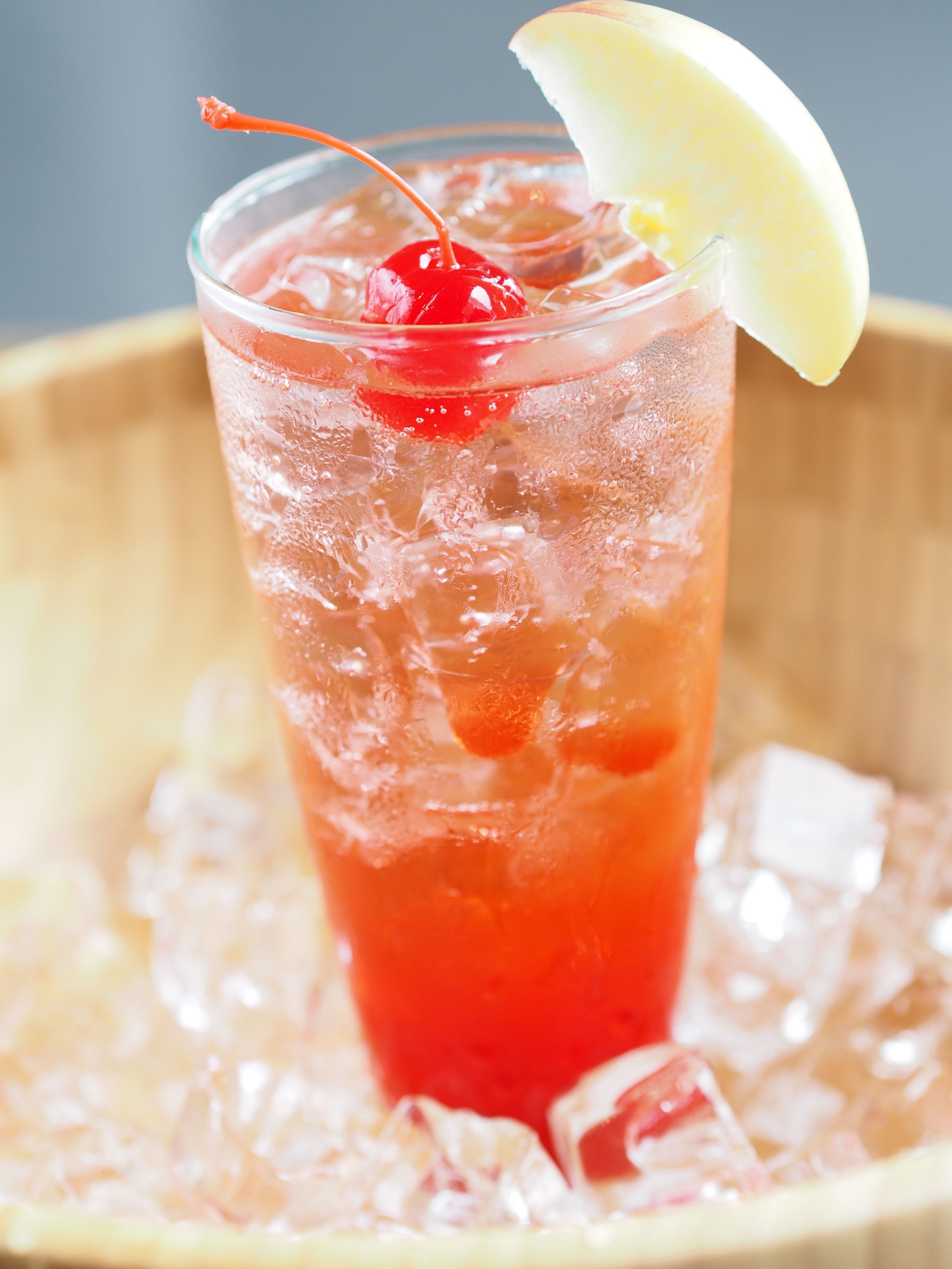 Un vaso de bebida roja con una cereza y una rodaja de limón, rodeado de cubitos de hielo.