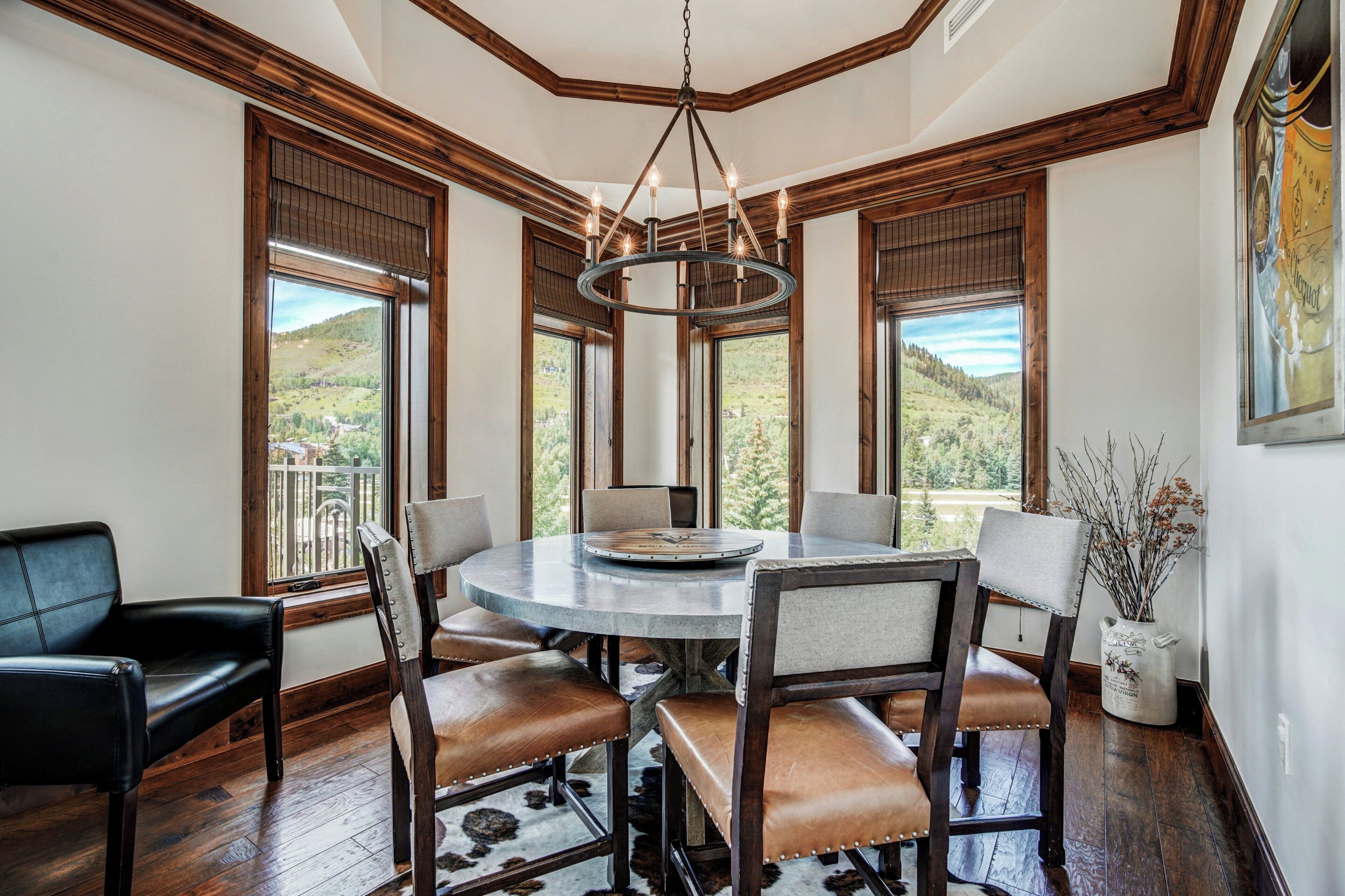 Uma sala de jantar com uma mesa redonda, seis cadeiras e um candelabro. Janelas amplas proporcionam vistas de uma paisagem montanhosa.