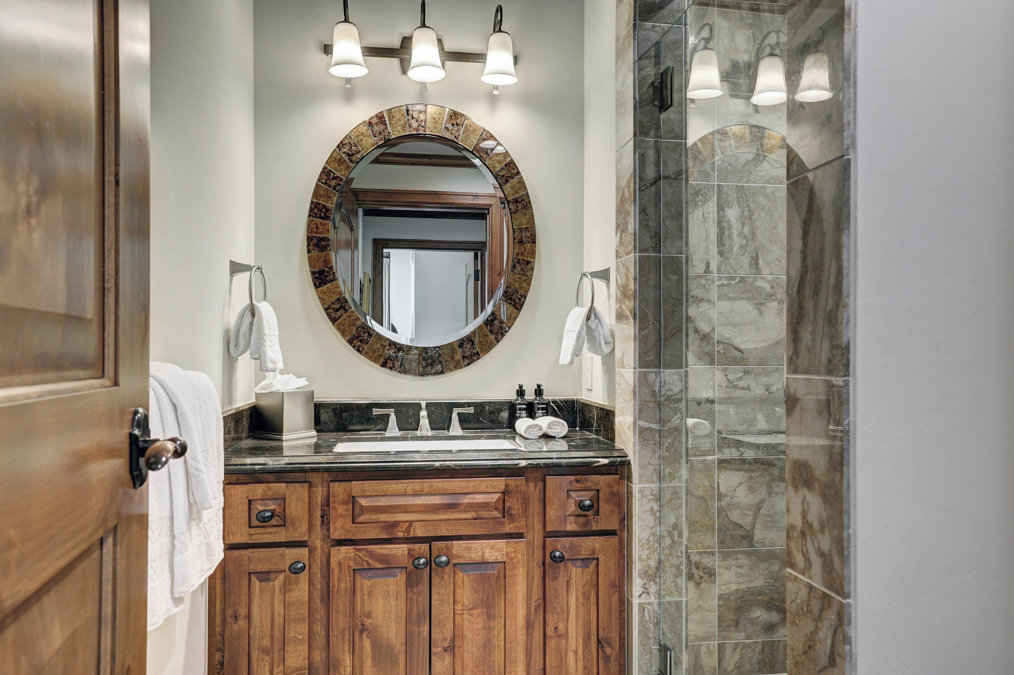 A bathroom with a wooden vanity, a round mirror with a stone frame, a sink with two faucets, and a shower with stone tiles.