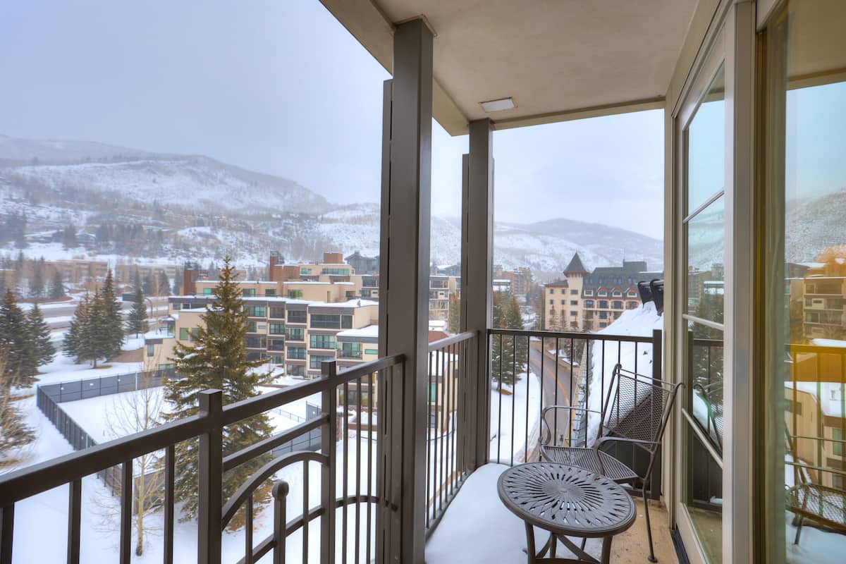 A balcony with a metal railing, a table, and two chairs. The balcony overlooks a snowy landscape with buildings and trees.