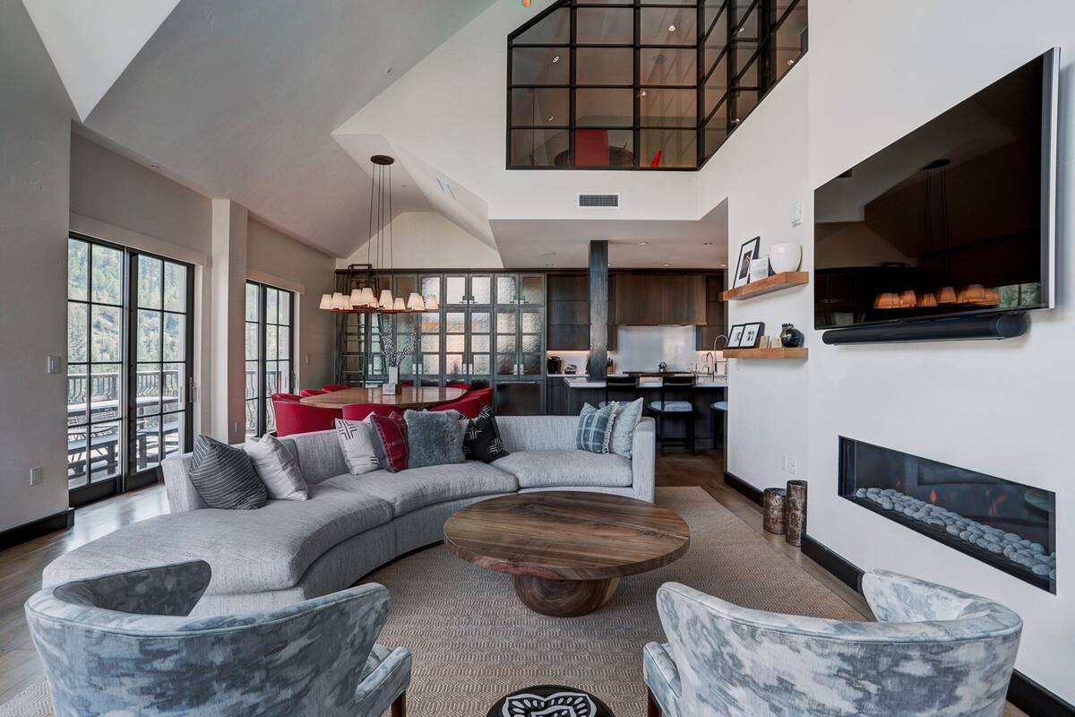 A spacious living room with a large sectional sofa, a wooden coffee table, and marble chairs. There is a fireplace, a kitchen area in the background, and large windows providing natural light.