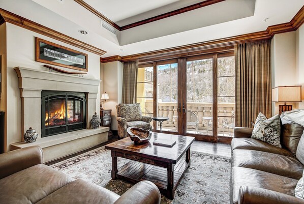 A living room with a fireplace, leather sofas, a coffee table, and a view of a mountain landscape through large windows.