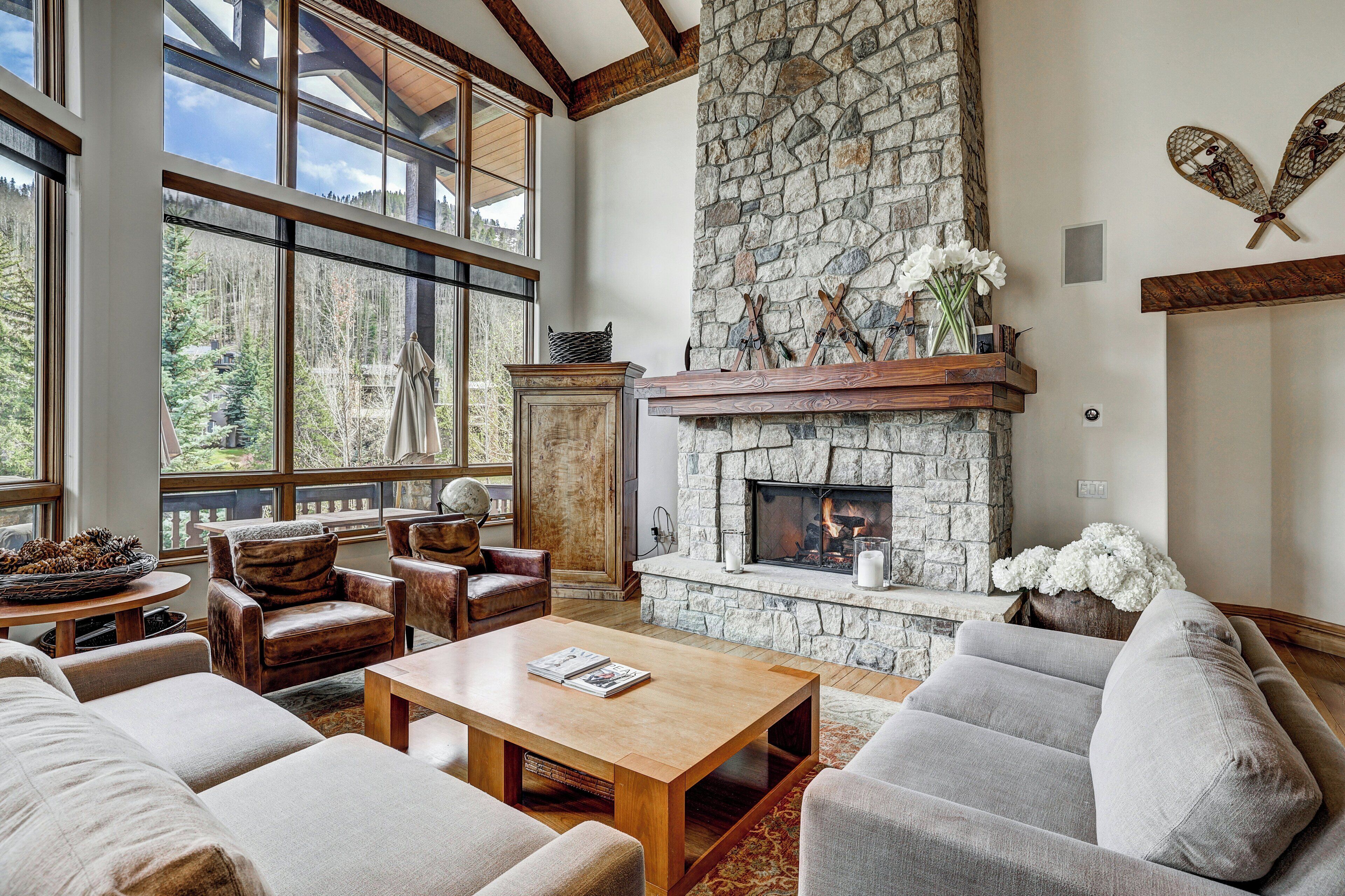 A spacious living room with a stone fireplace, leather sofas, and a large window offering a view of the forest.