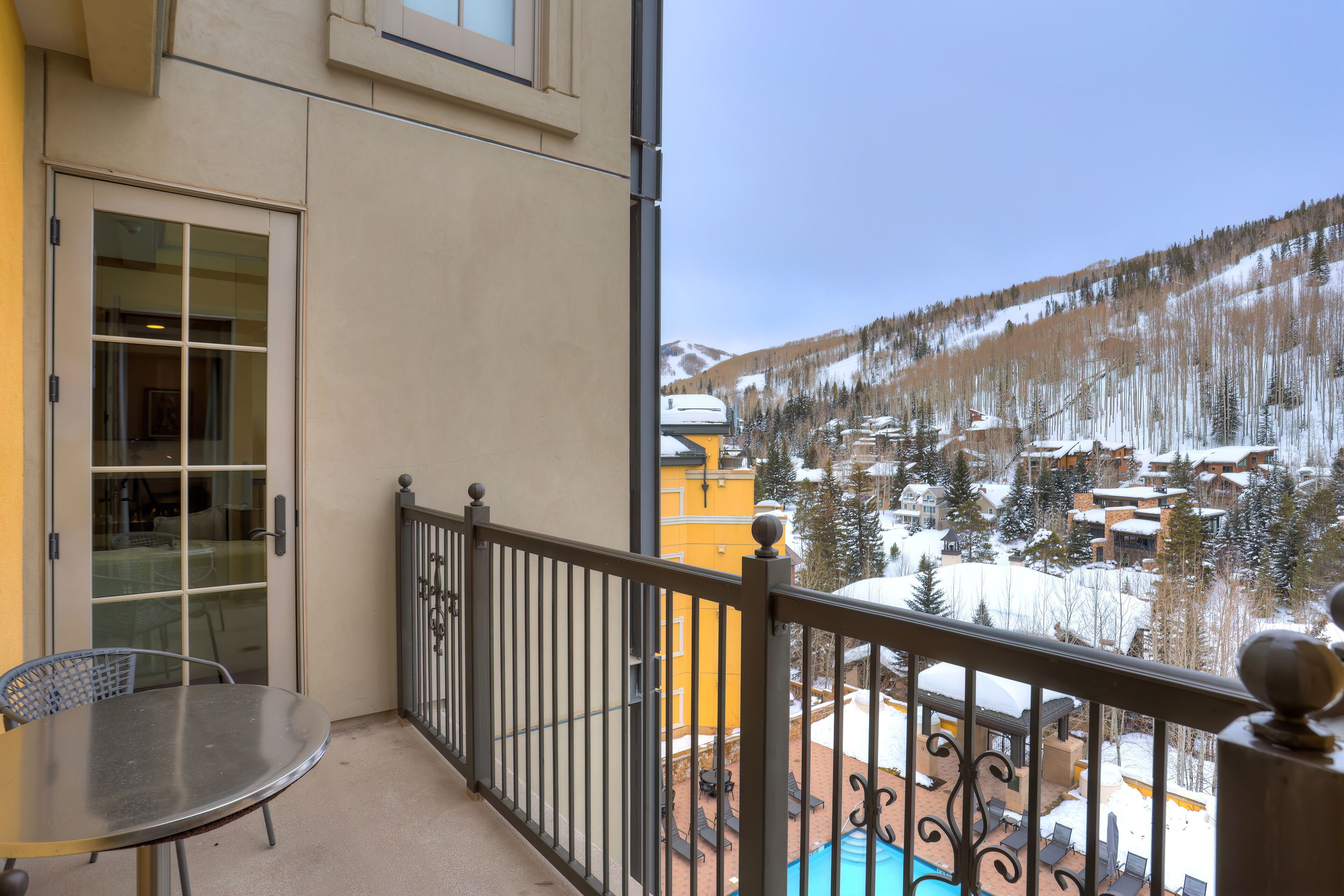 A balcony with a glass door, a table, and chairs, overlooking a snowy mountain landscape with buildings and trees.