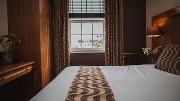 A hotel room with a large bed, wooden wardrobe, a window with curtains, a desk with a vase of flowers, and a lamp on the nightstand.