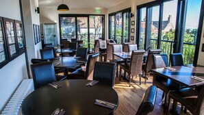 Un restaurant avec des tables rondes et des chaises noires, de grandes fenêtres donnant sur des immeubles et des arbres, et un luminaire au plafond.