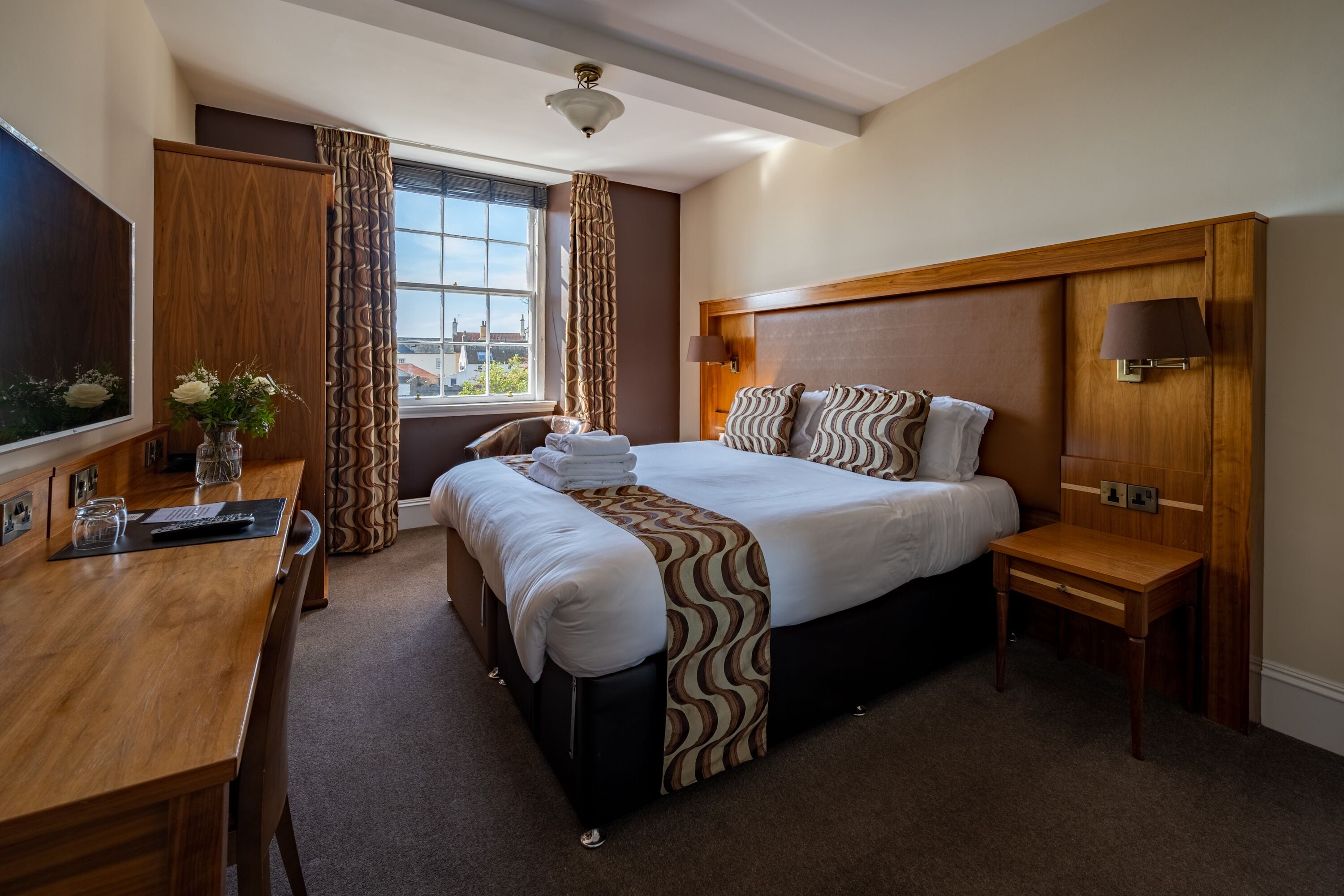 A hotel room with a large bed, a desk, a chair, a TV, a window with curtains, and a lamp.