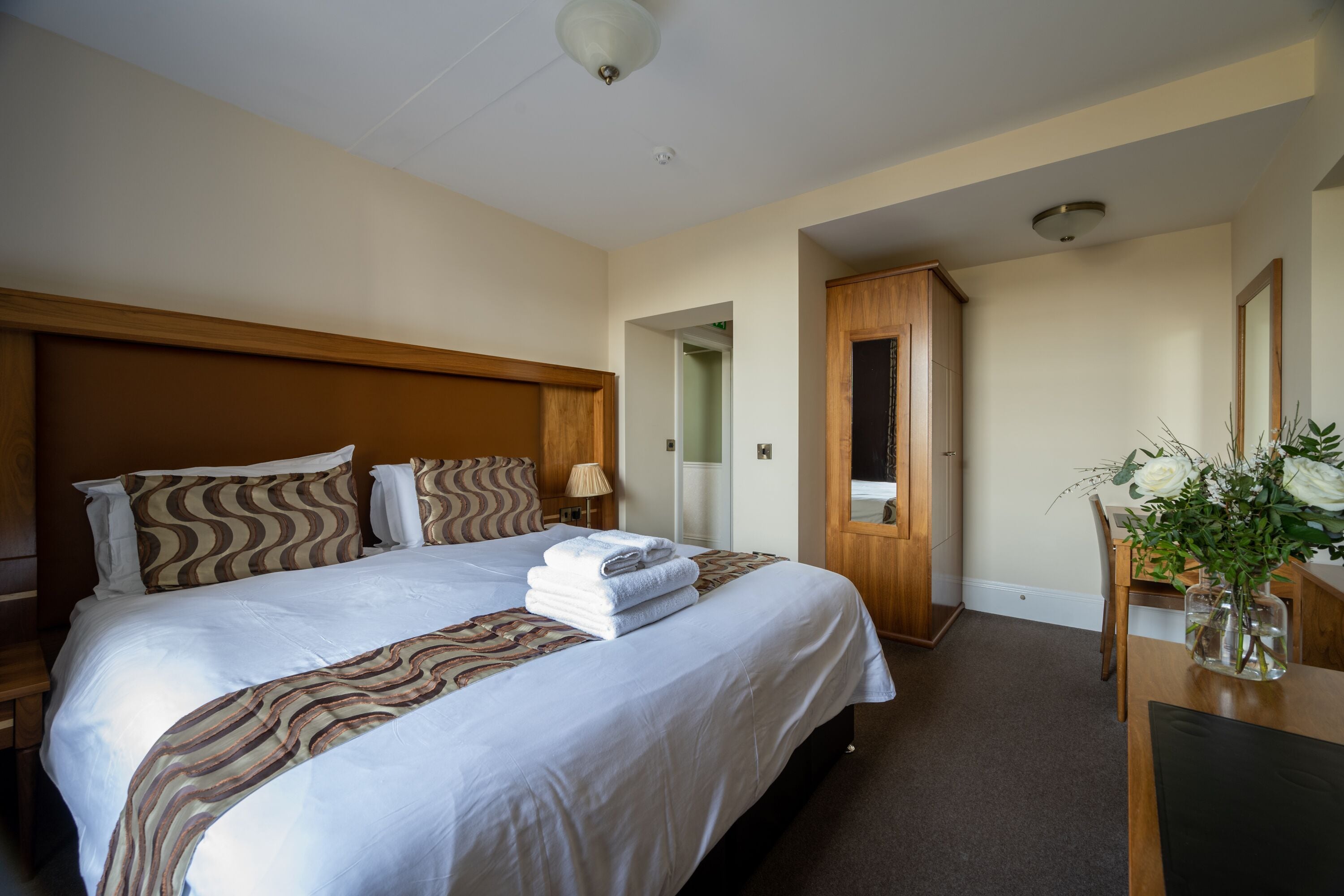 A hotel room with a large bed, wooden headboard, a desk with a vase of flowers, and a door leading to another room.