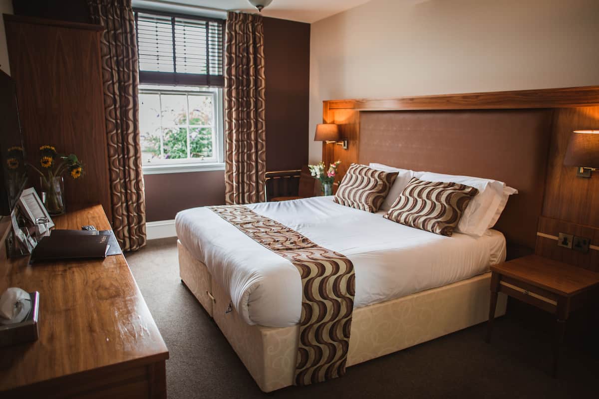 A hotel room with a large bed, wooden headboard, a desk with a lamp, and a window with curtains.