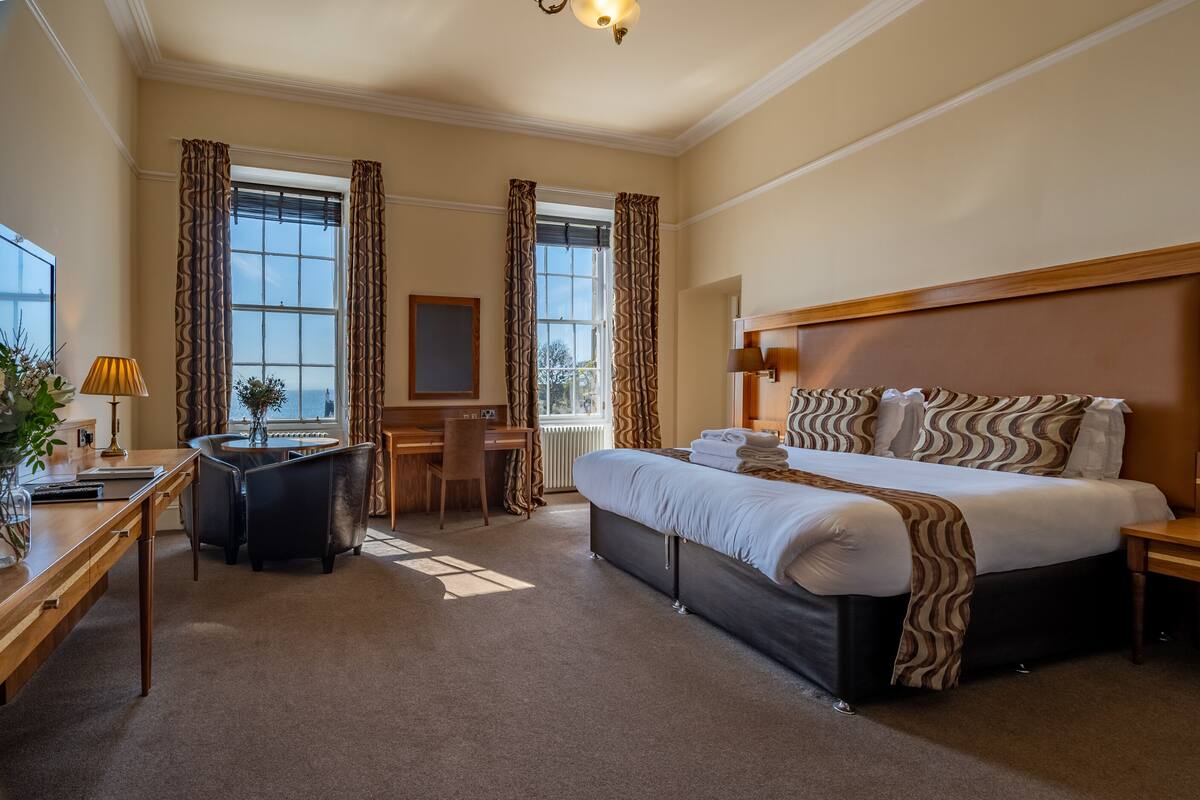 A hotel room with a large bed, a desk with a chair, a small table, and a window with curtains.