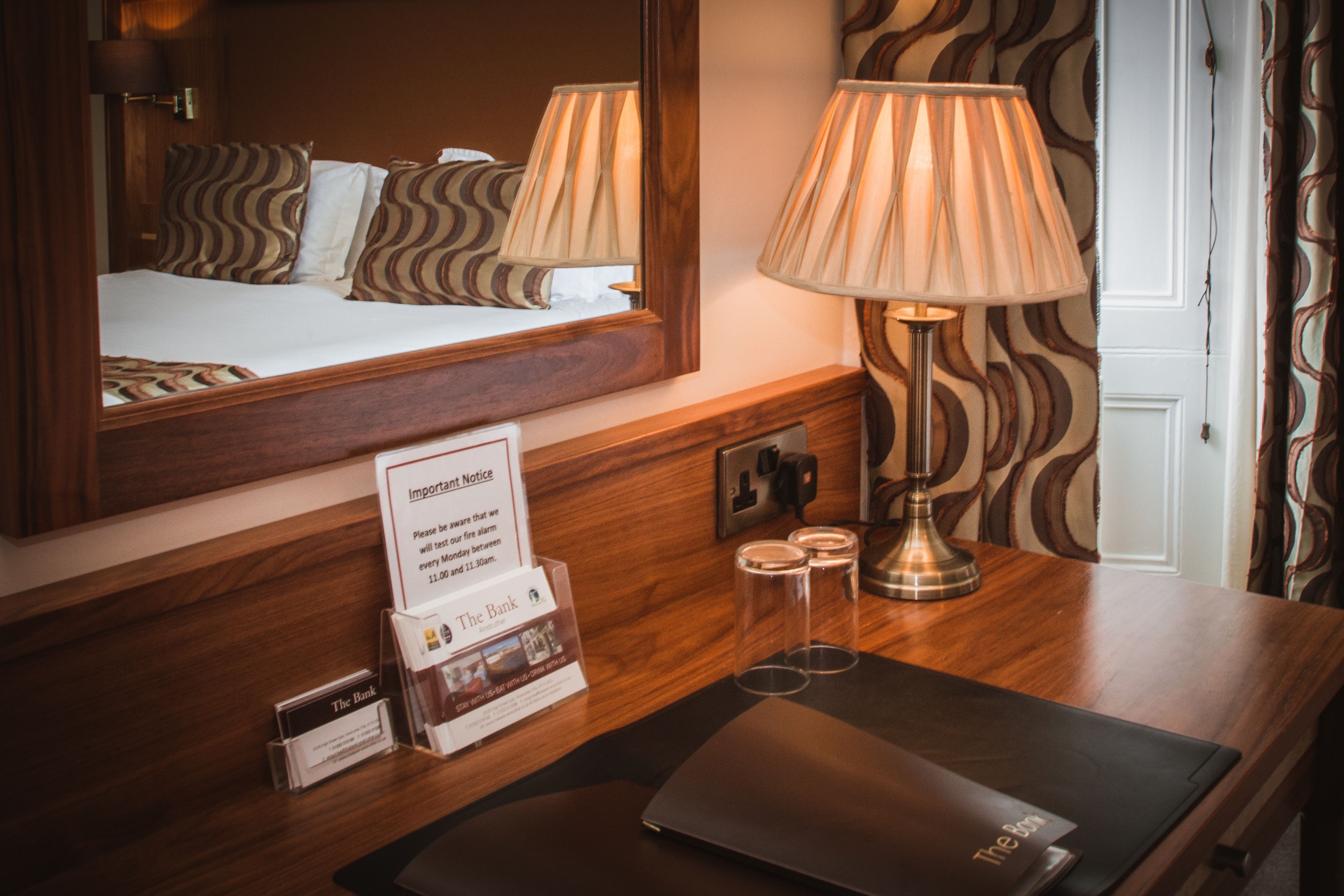 Habitación de hotel con cama, lámpara de noche, escritorio con una laptop y un espejo que refleja la habitación. Hay un cartel en el escritorio y una puerta con cortina estampada.