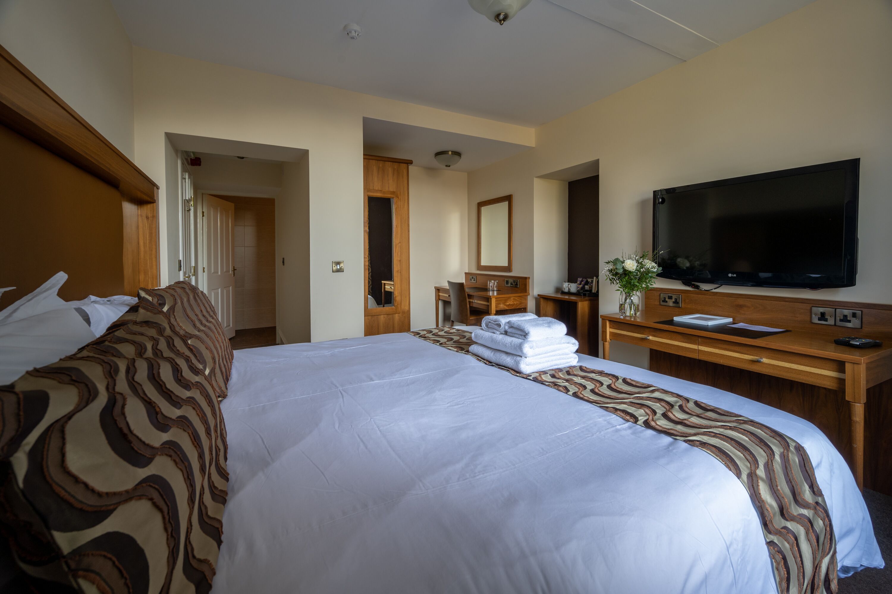 A hotel room with a large bed, a wooden desk, a flat-screen TV, and a bathroom visible through an open door.