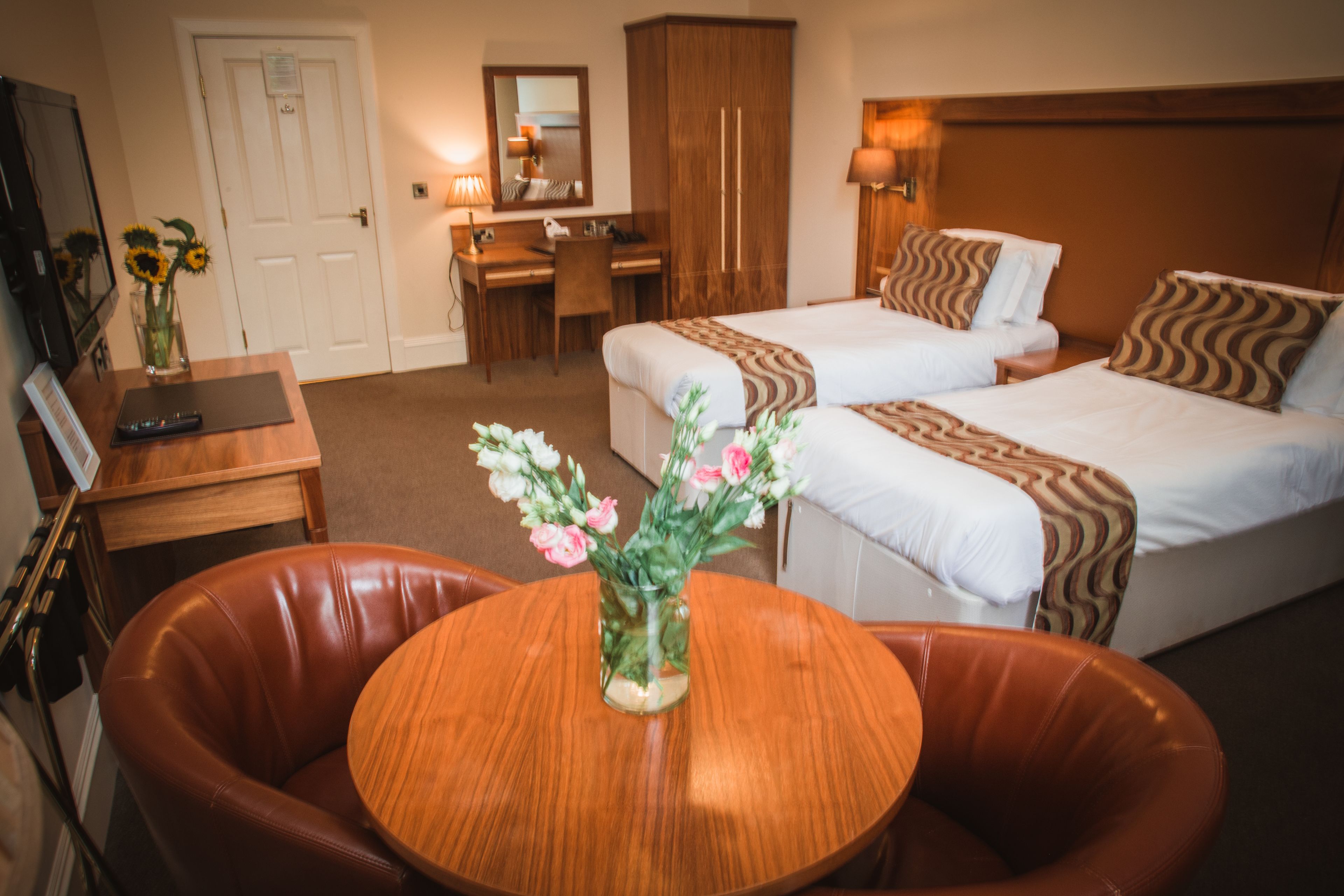 A hotel room with two beds, a desk, a chair, and a round table with a vase of flowers.