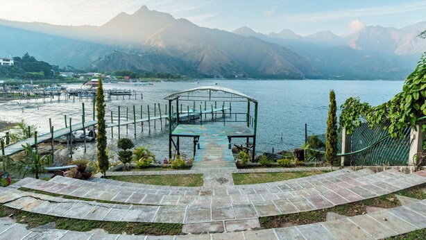 A lakeside walkway leading to a covered seating area with a view of mountains and boats.
