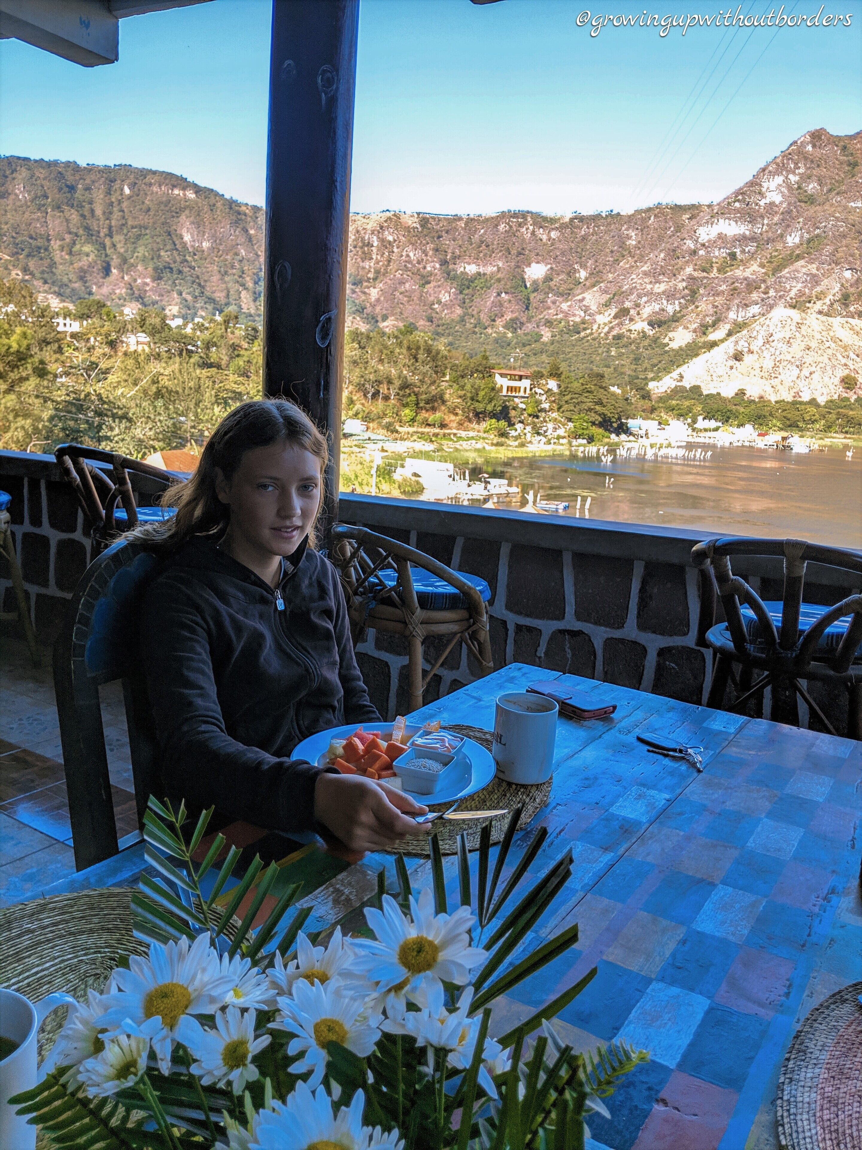 A person seated at a table with a view of a river and mountains in the background.