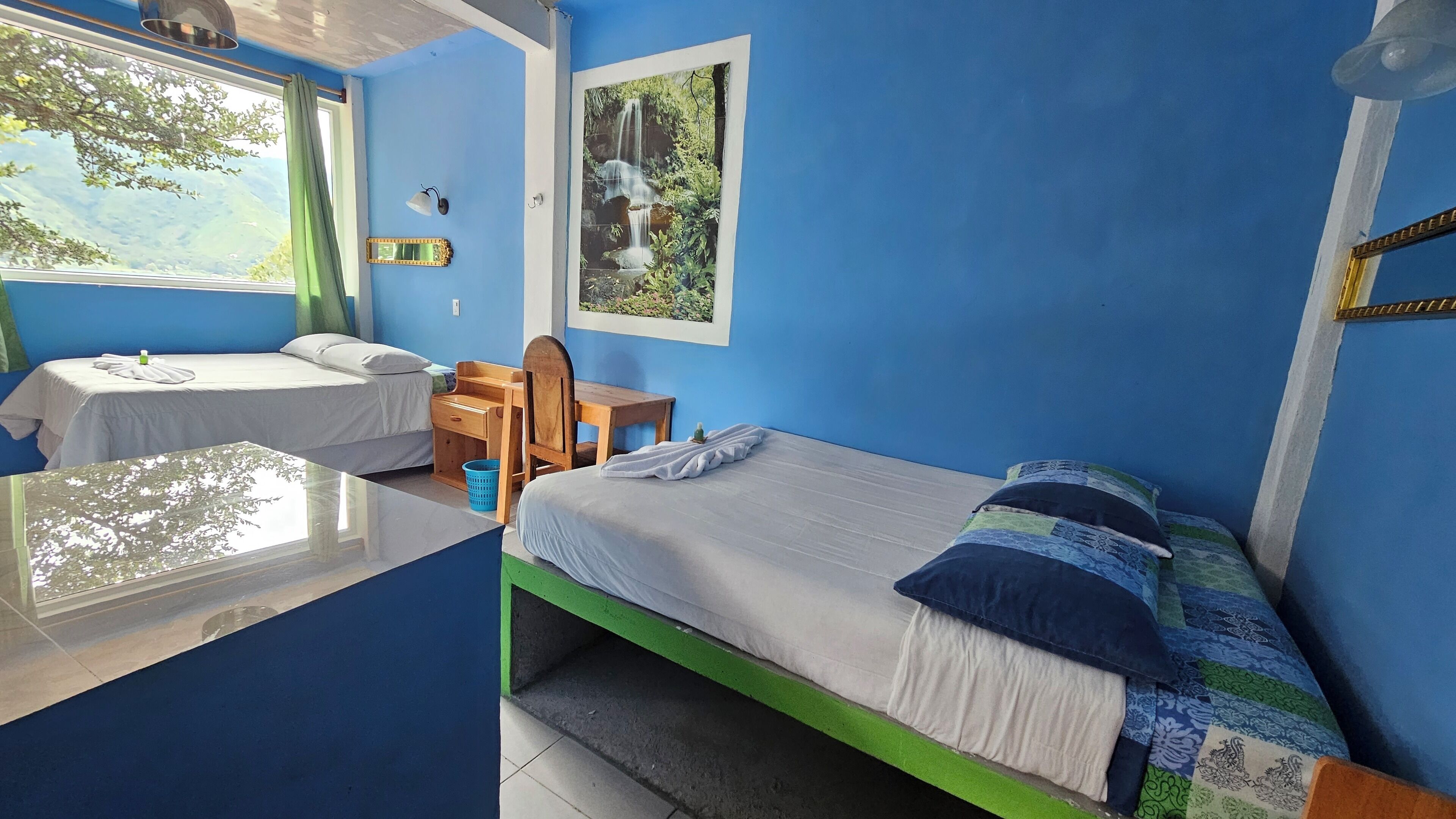 Una habitación con dos camas, una mesita, una silla, un ventanal con vistas a zonas verdes y un cuadro en la pared.