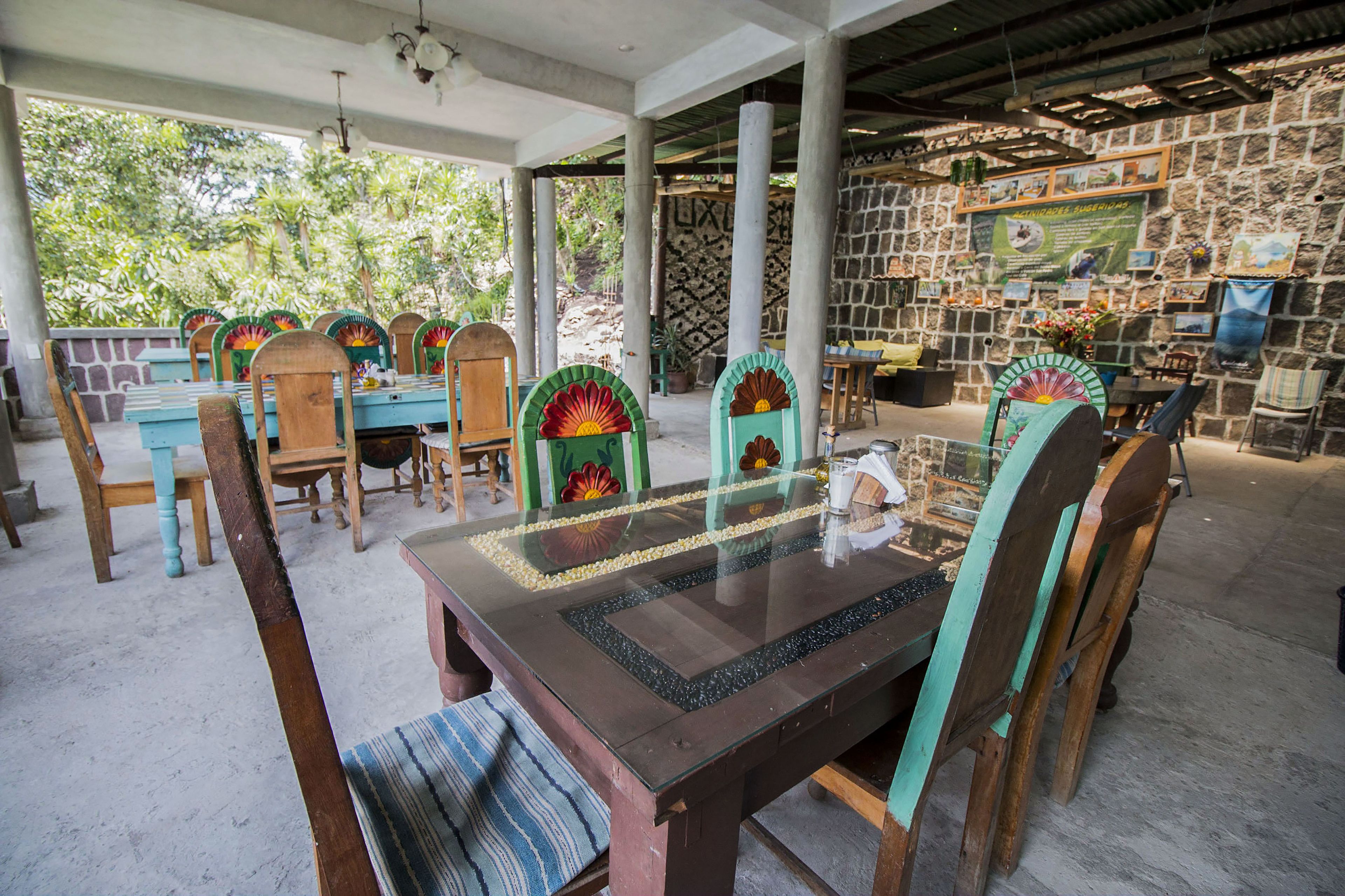 Un espacio comedor al aire libre cubierto, con pared de piedra, sillas de madera y mesa con superficie de vidrio.