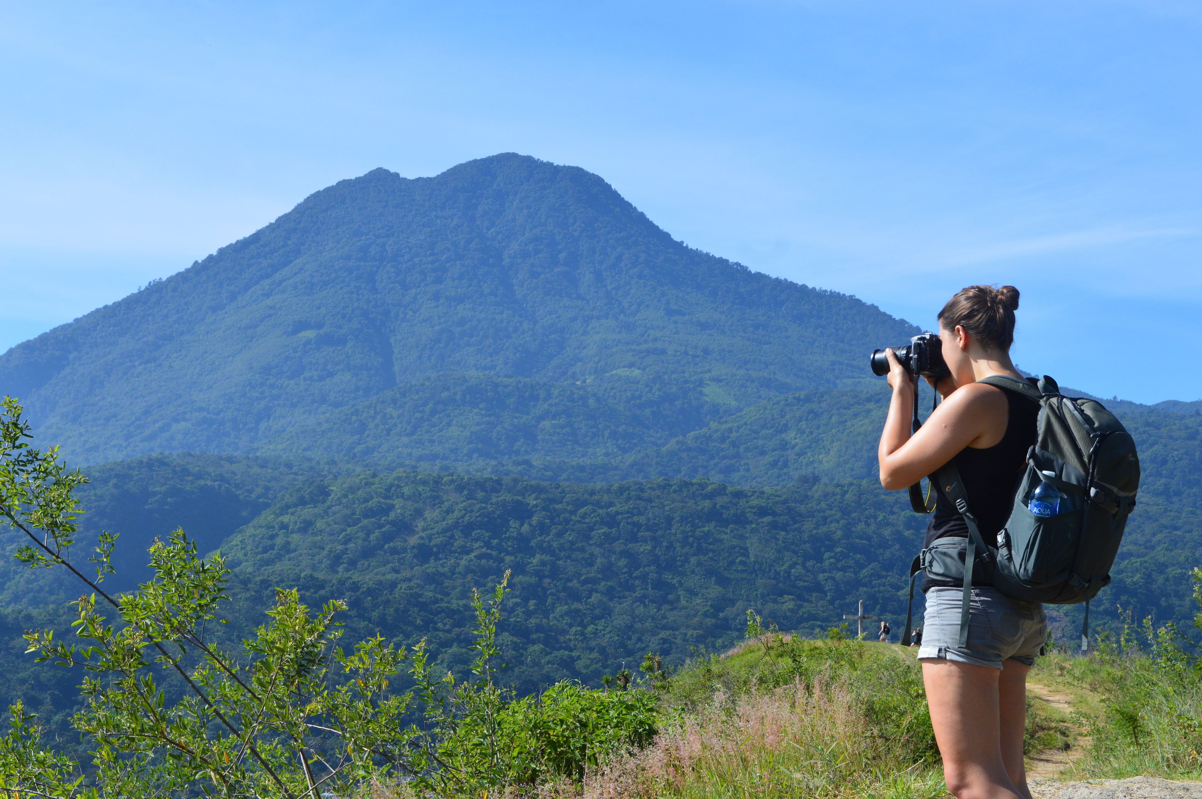 A person with a camera and a backpack standing on a path, overlooking a mountainous landscape.