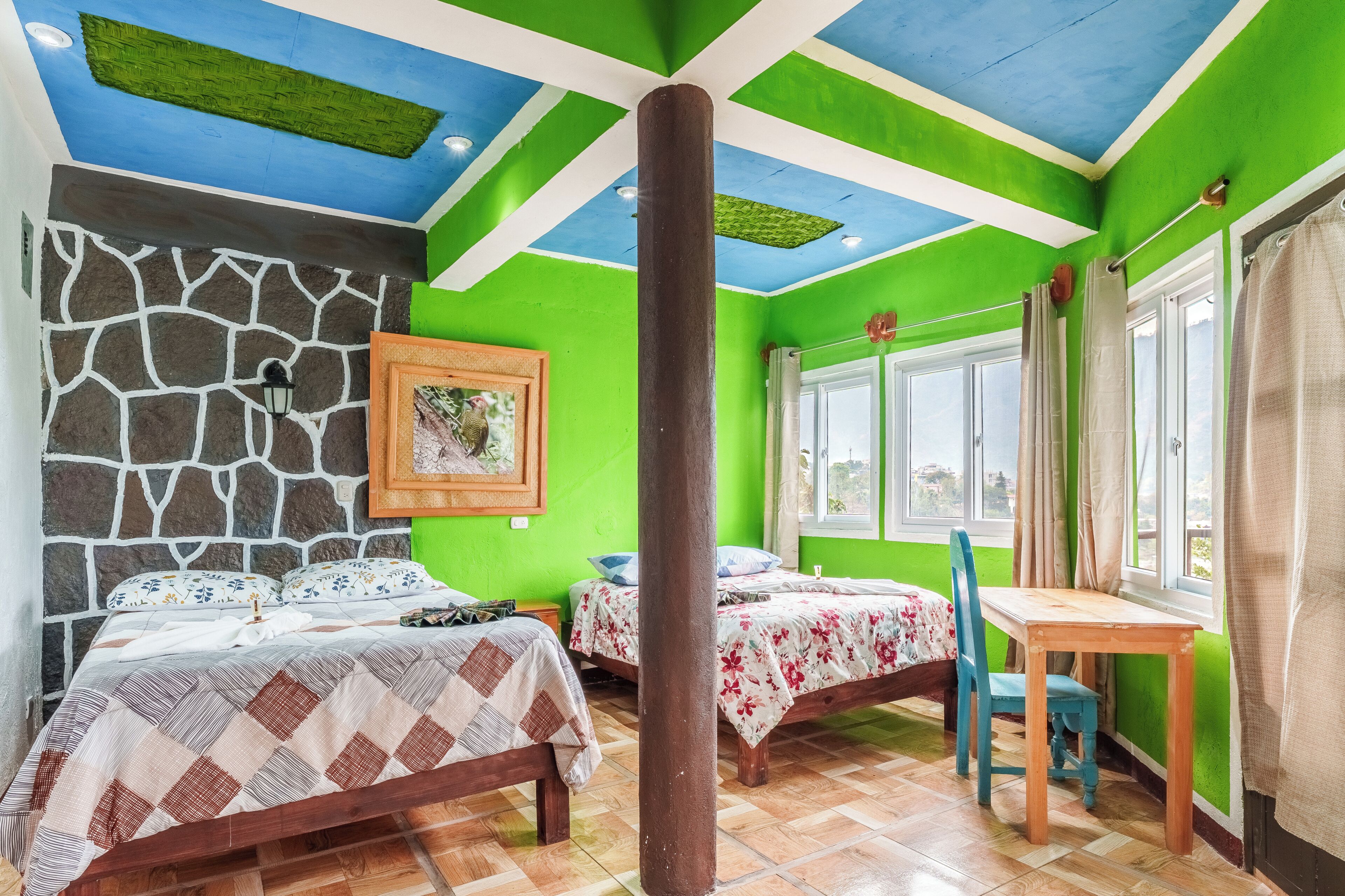 Una habitación con dos camas, una mesa de madera, una pared de piedra y una ventana con cortinas.