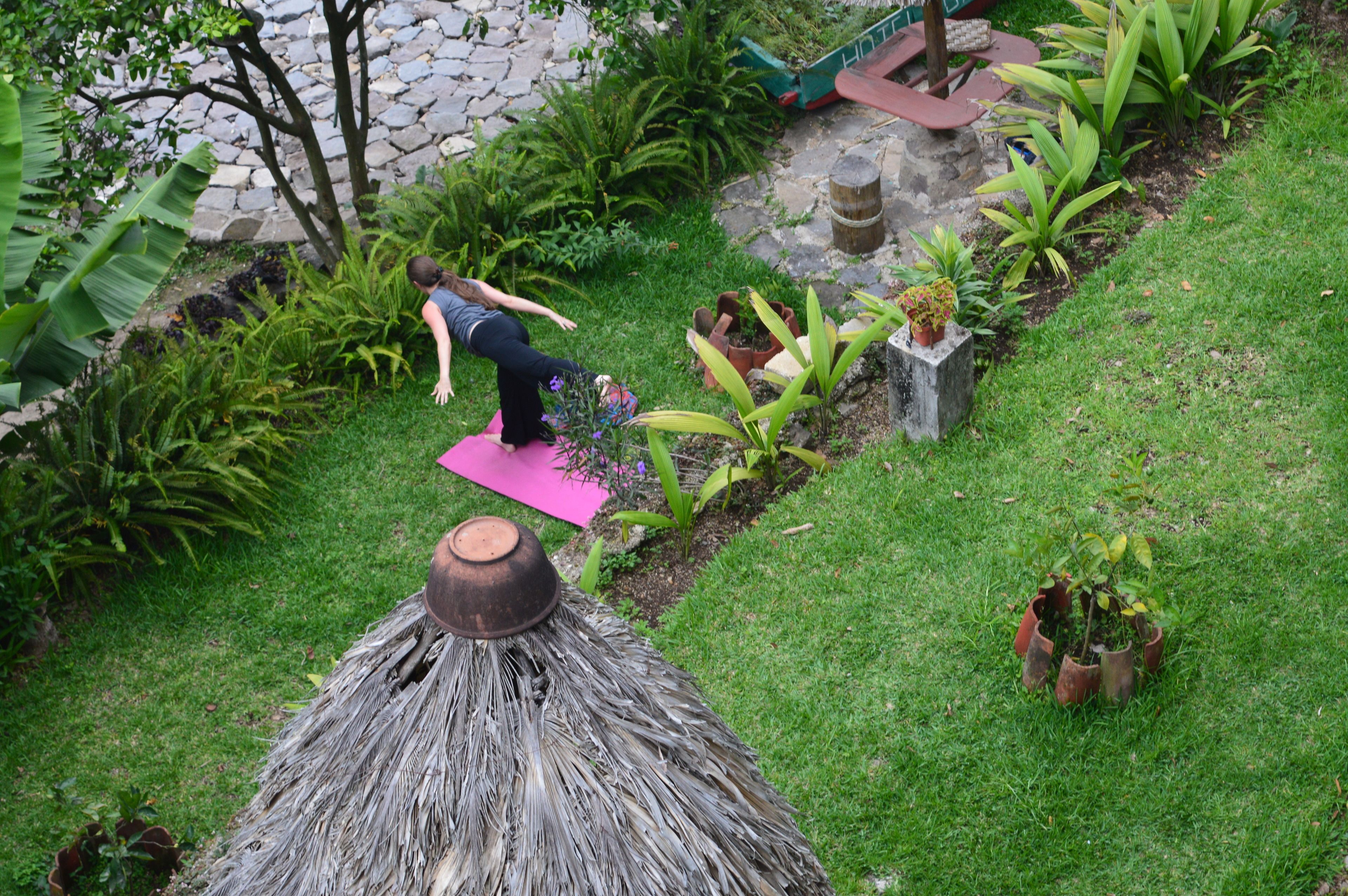 Une personne pratiquant le yoga sur un tapis dans un jardin, entourée de différentes plantes et d’une structure à toit de chaume.