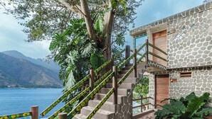 A stone staircase leading to a building with a wooden door, surrounded by greenery and overlooking a body of water.