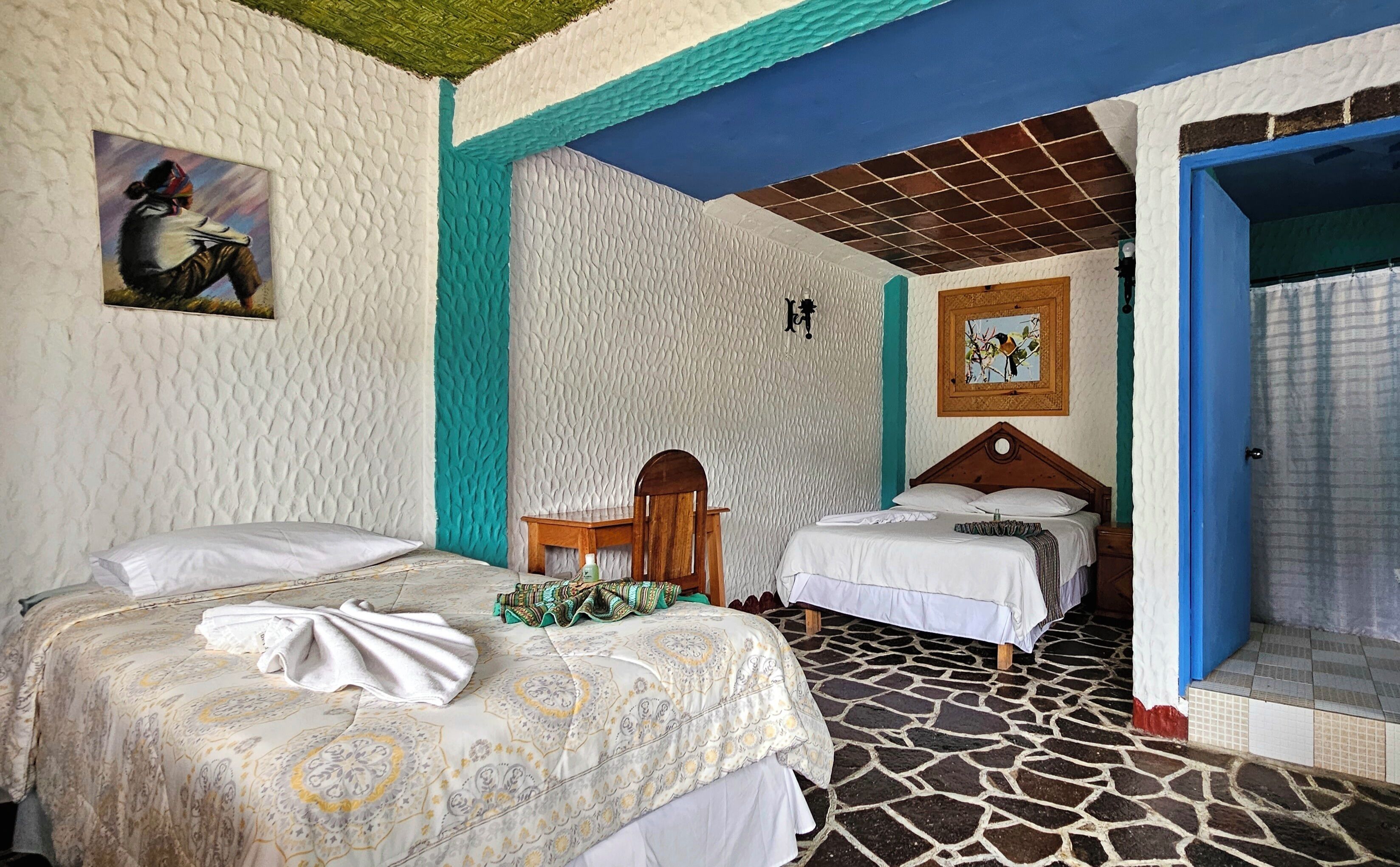 Una habitación con dos camas, una silla de madera, una mesita pequeña y un baño con ducha.
