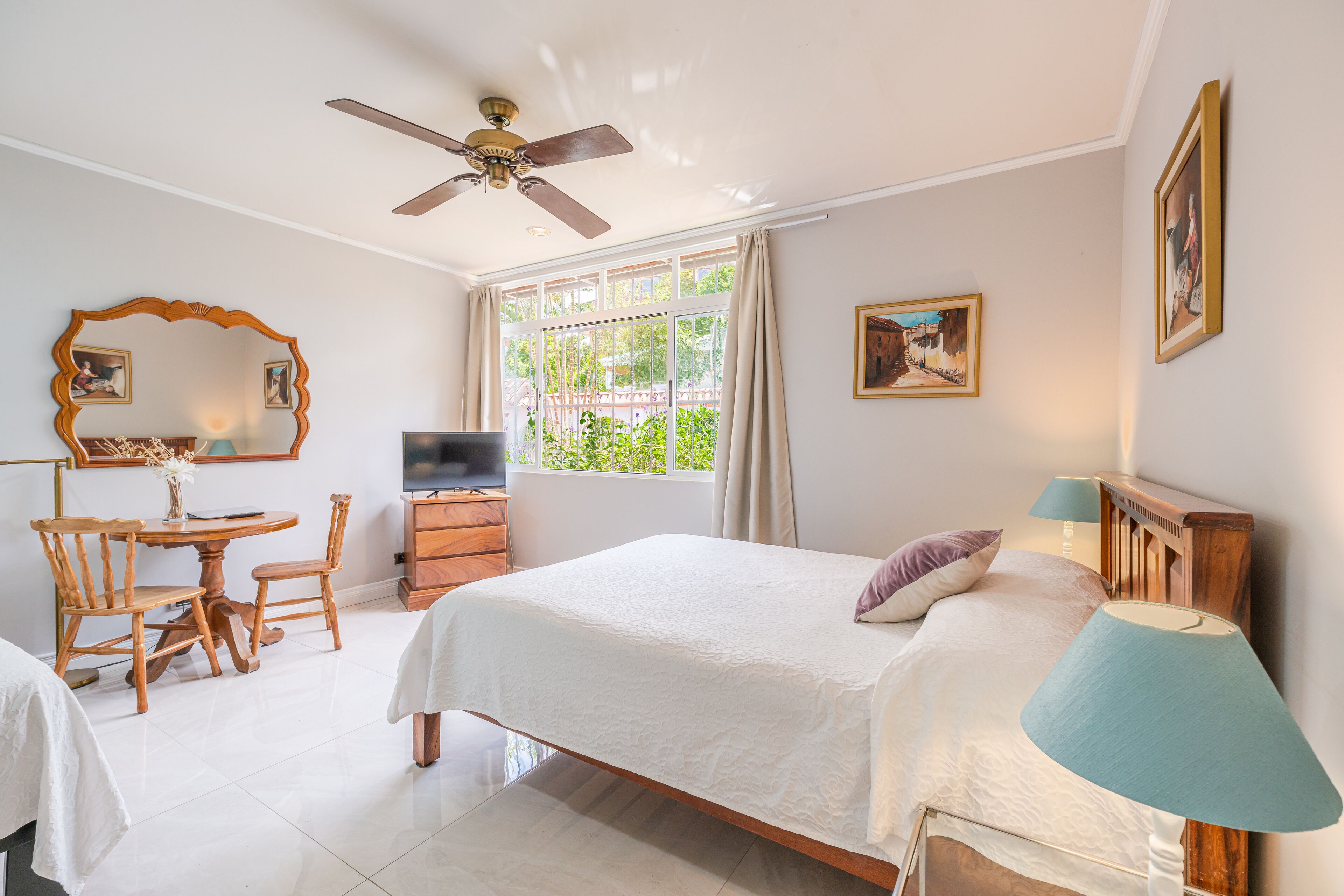 A hotel room with a bed, a desk with a chair, a TV, a ceiling fan, and a window with curtains.