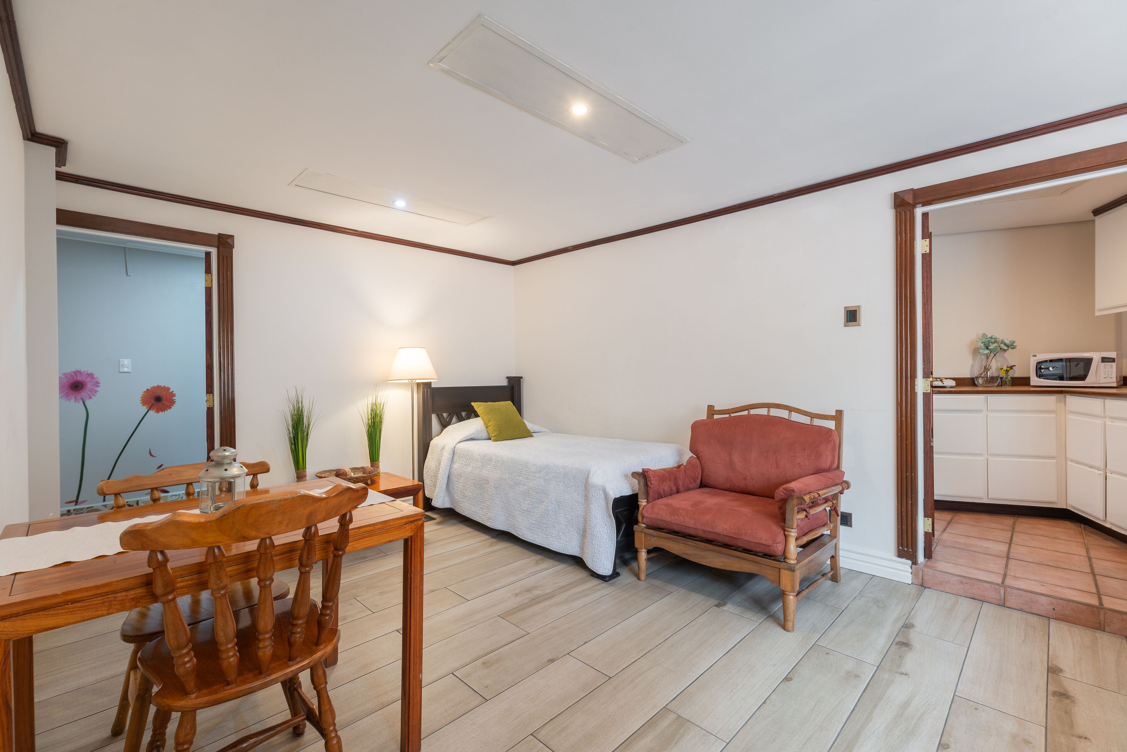 Ein Zimmer mit einem Bett, einem roten Sessel, einem Holzespeisetisch mit Stühlen, einer Lampe und einer Küchenzeile.