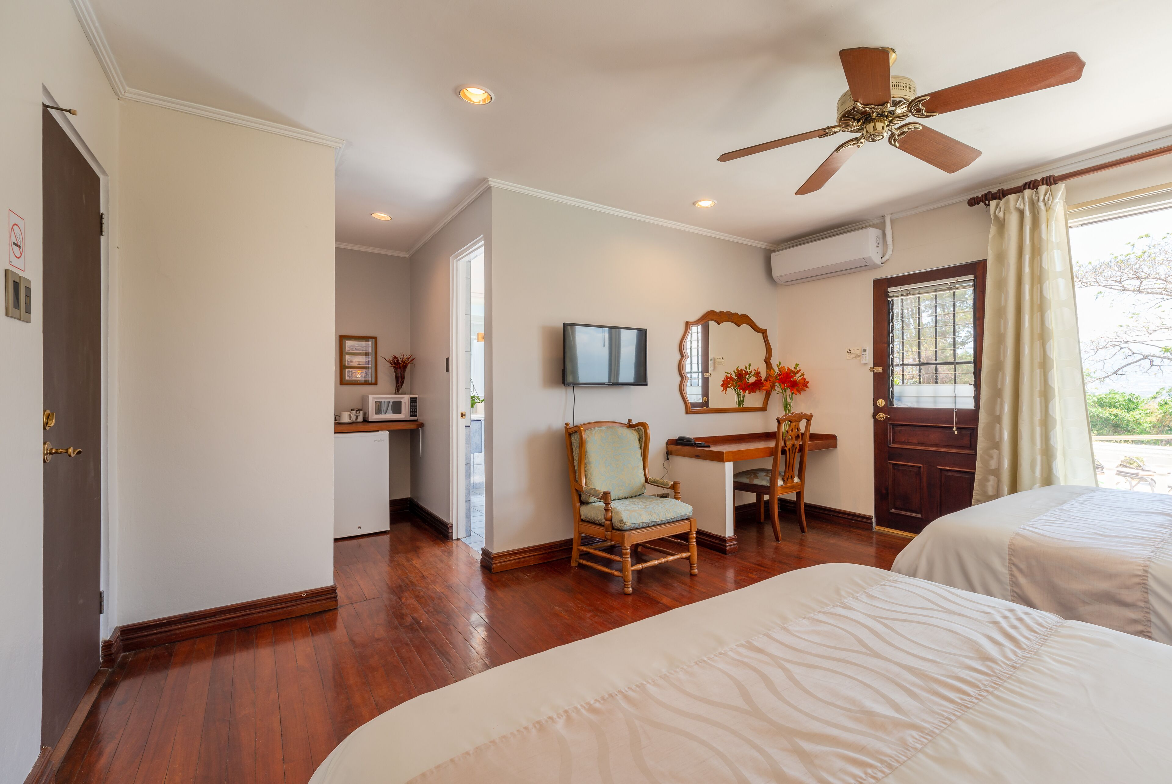 A hotel room with a wooden floor, a ceiling fan, a desk with a chair, a TV, and a bed.