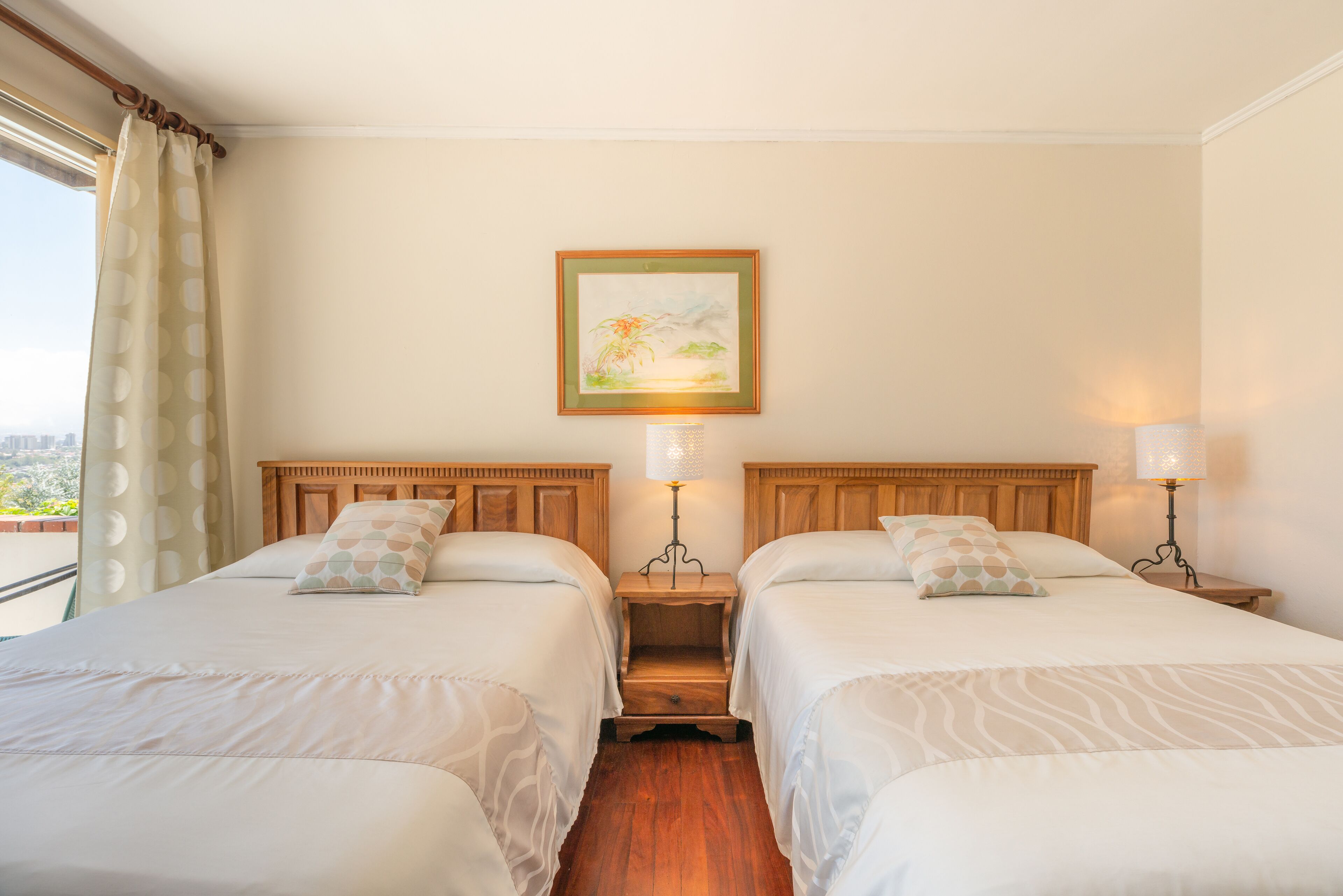 A hotel room with two beds, a nightstand with lamps, a painting on the wall, and a view through the window.