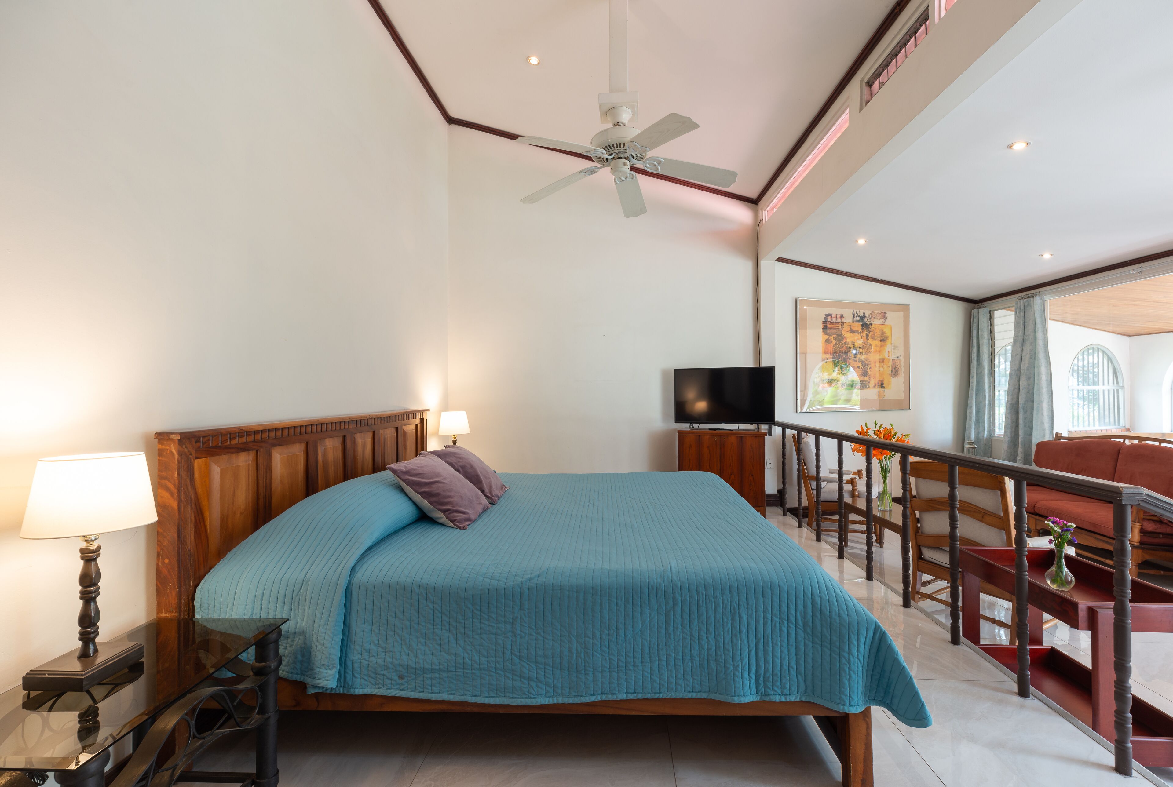 A spacious bedroom with a large bed, a ceiling fan, a TV, and a balcony with a view.