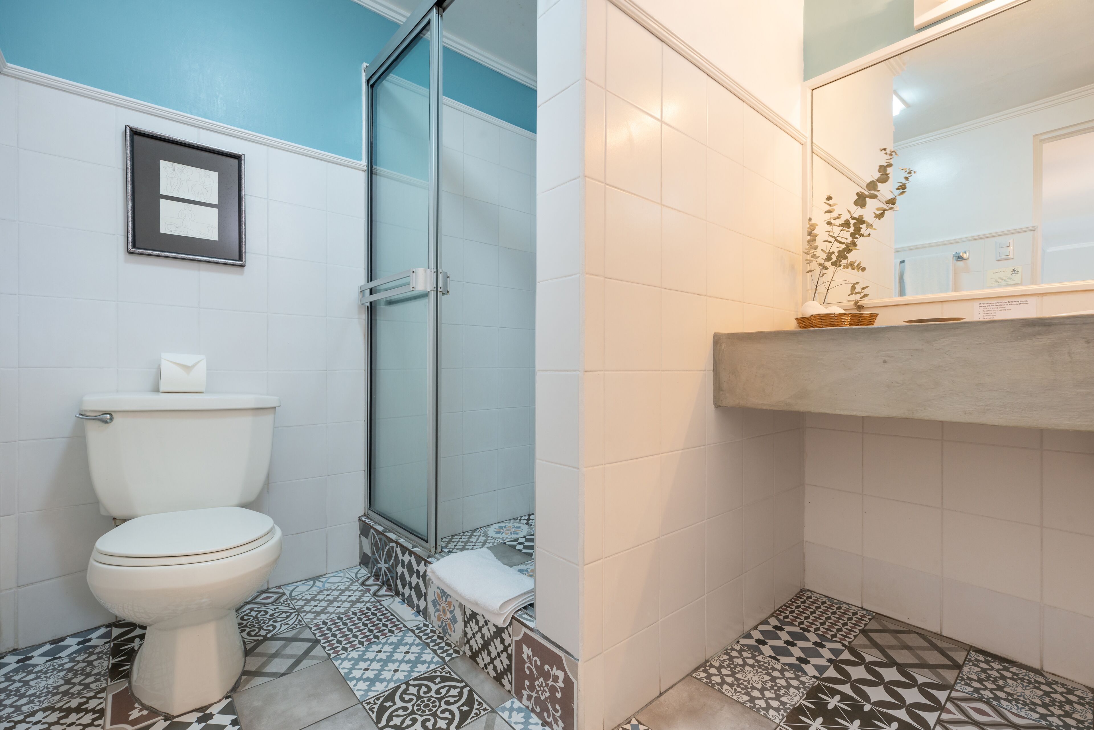 A bathroom with a toilet, shower, tiled floor, and a mirror with a plant.