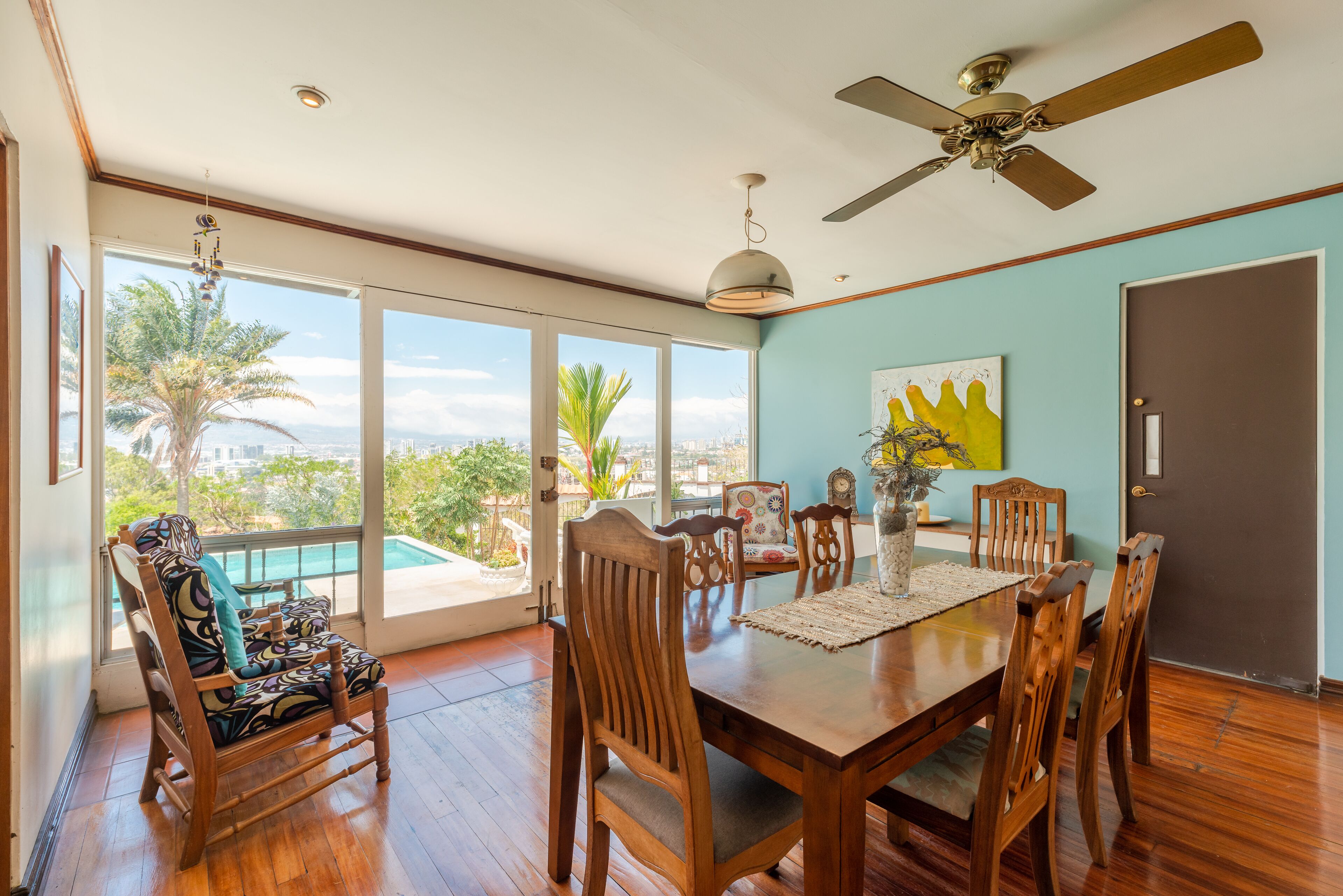 A dining area with a wooden table, chairs, and a view of palm trees and a cityscape through large windows.